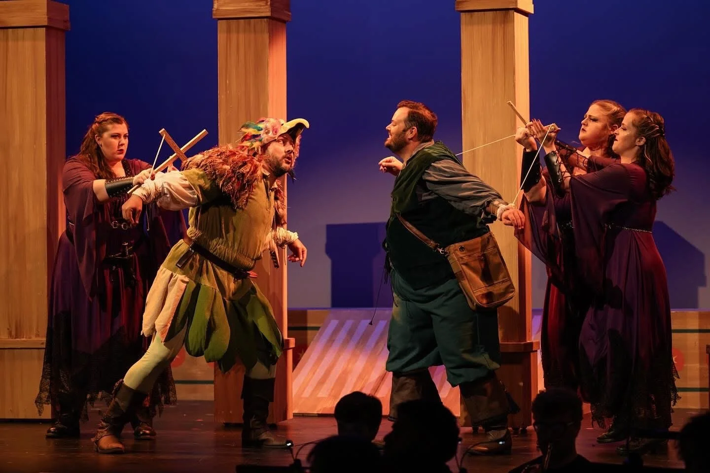 Theatrical scene with five actors in costume on stage, portraying characters in a struggle or confrontation, with a simplistic wooden set background.