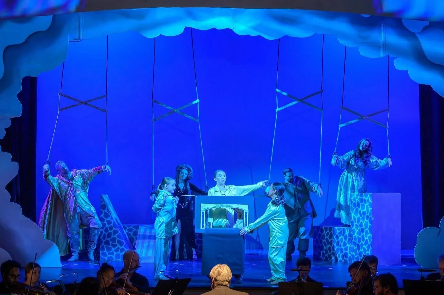 Children's theater performance on stage with blue background, puppets, and actors in costumes, with orchestra in front.