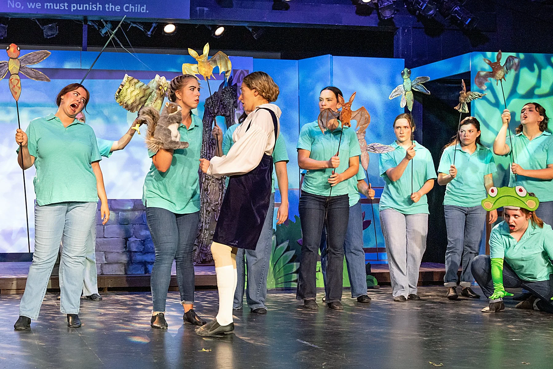 Stage scene from a play featuring women in green costumes holding puppet props of insects and animals, with a woman in a frog costume squatting on the right.