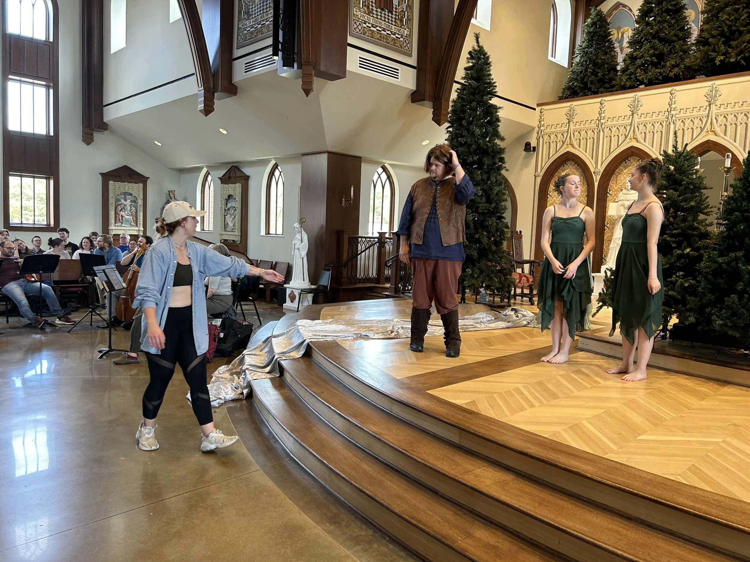 Actors rehearsing a play on a stage decorated with Christmas trees and gothic-style architecture, with an audience and a director present.