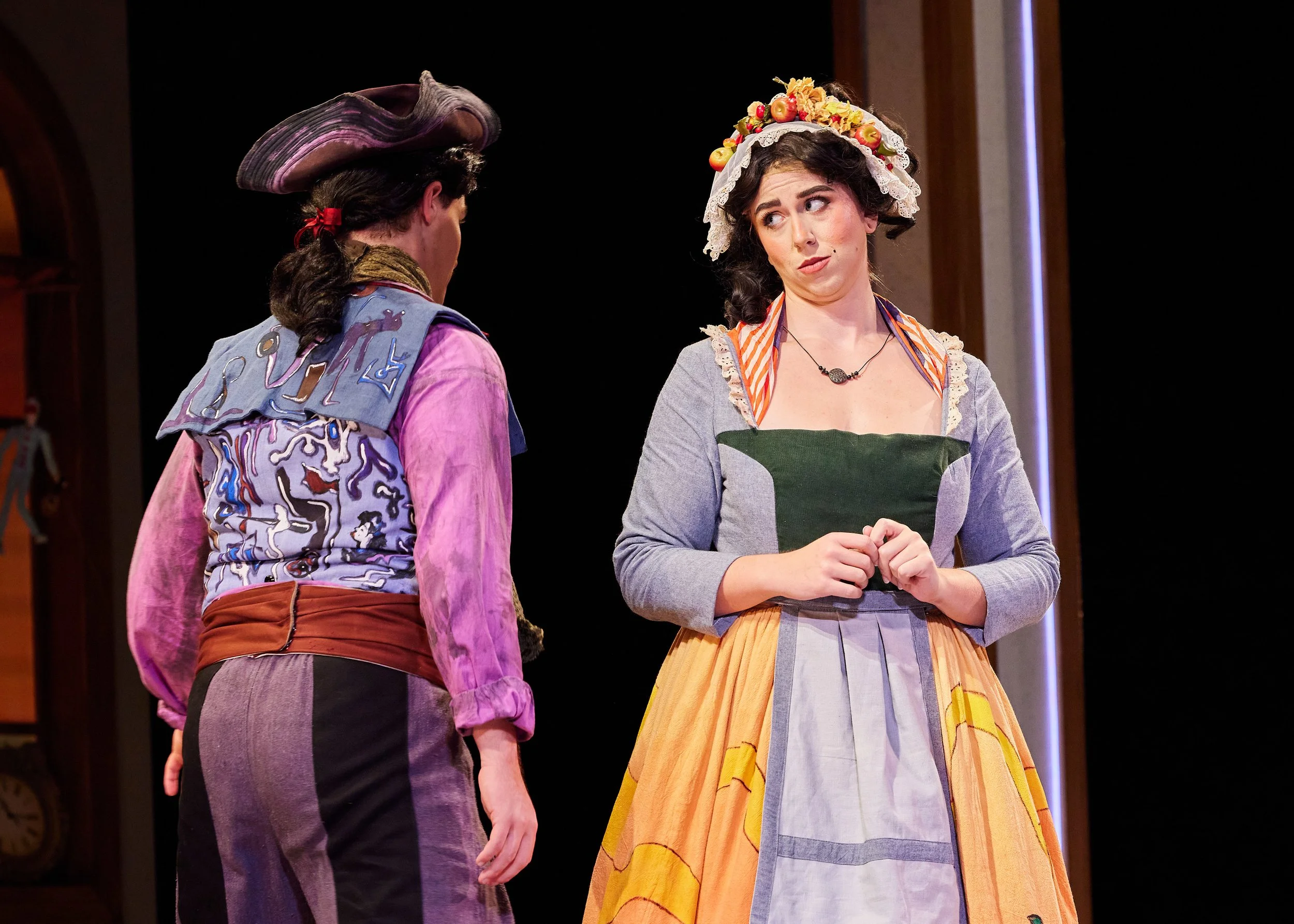 Two women talking on stage in period costumes, one wearing a hat decorated with fruit, the other wearing a patchwork vest and a large skirt.