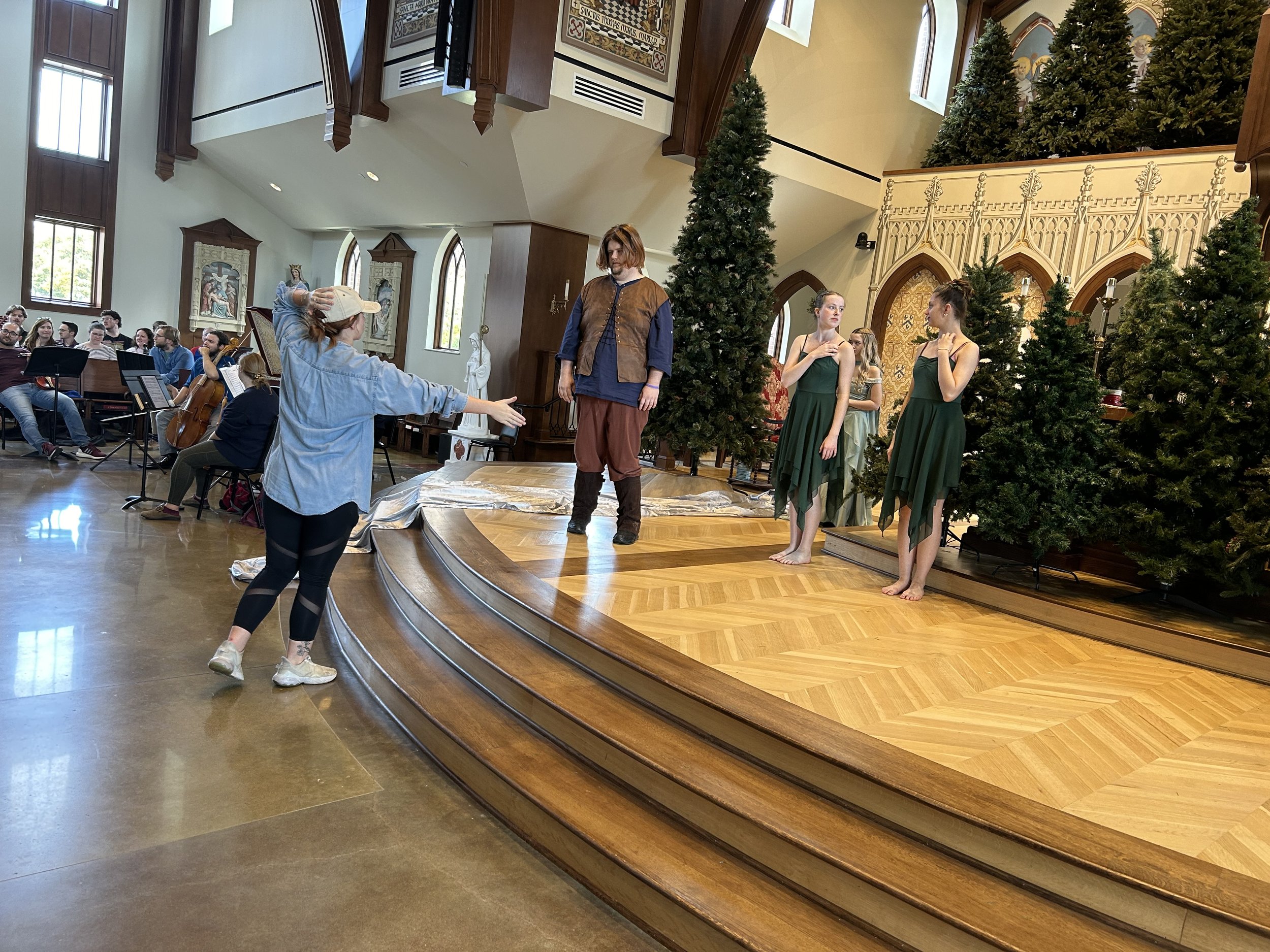 Director talking to three dancers in green costumes during a rehearsal on a wooden stage with Christmas trees, while an orchestra plays in the background inside a church or performance hall.