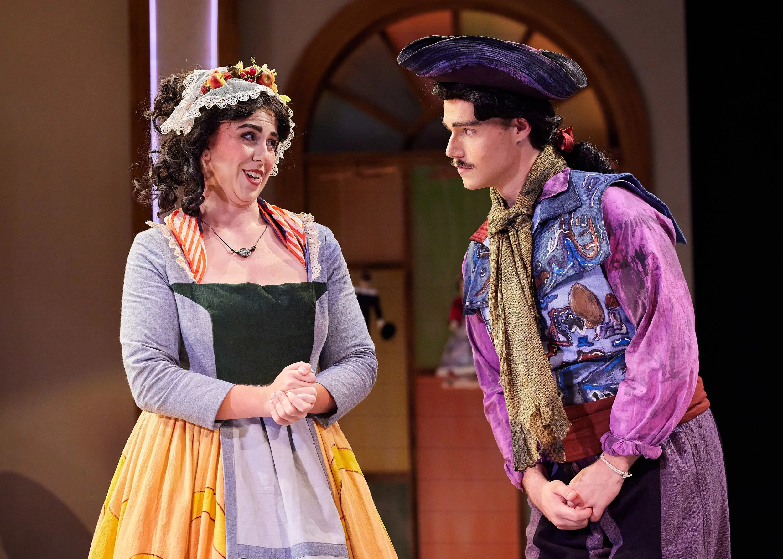 A woman in vintage dress with floral headpiece and a man in colorful costume and wide-brimmed hat engaged in conversation on stage.