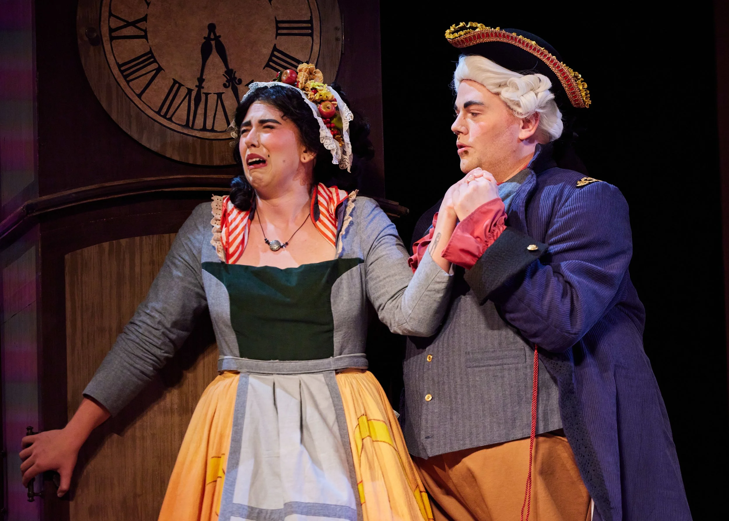 The image shows two actors on stage in costume. The woman on the left is crying and appears distressed, wearing a dress and apron, with a floral headpiece. The man on the right is in colonial military attire, holding the woman's hand, appearing serio