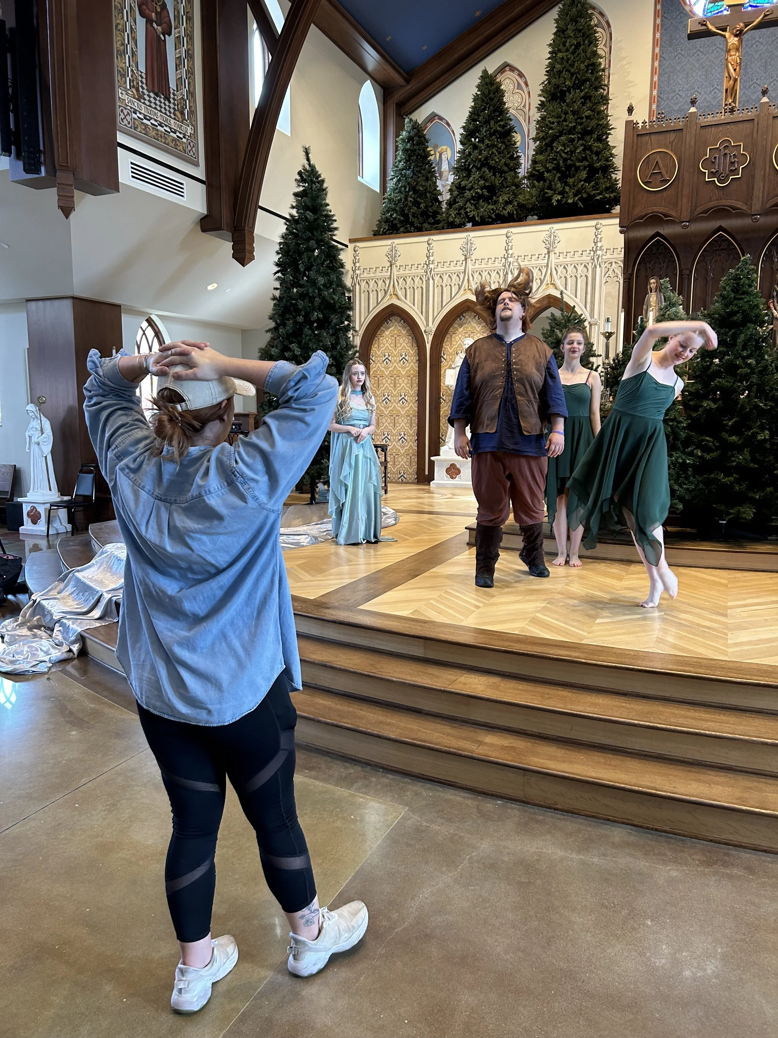 A woman is taking a photo of a theatrical performance on stage inside a church or cathedral. The stage has four performers dressed in costumes, with three women in green dresses and one man with long hair in fantasy-style clothing. The stage backdrop