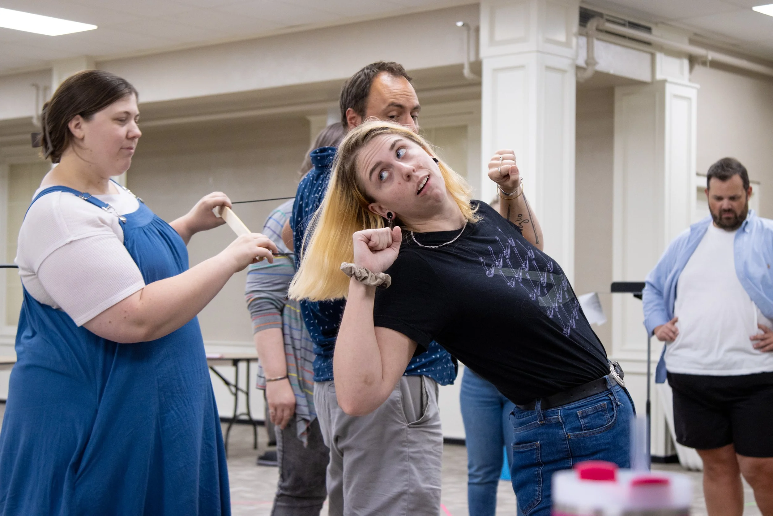 A woman with blonde hair and a black shirt appears to be playfully pulling a string attached to her head or a person's head behind her, while others observe in the background inside a large room.