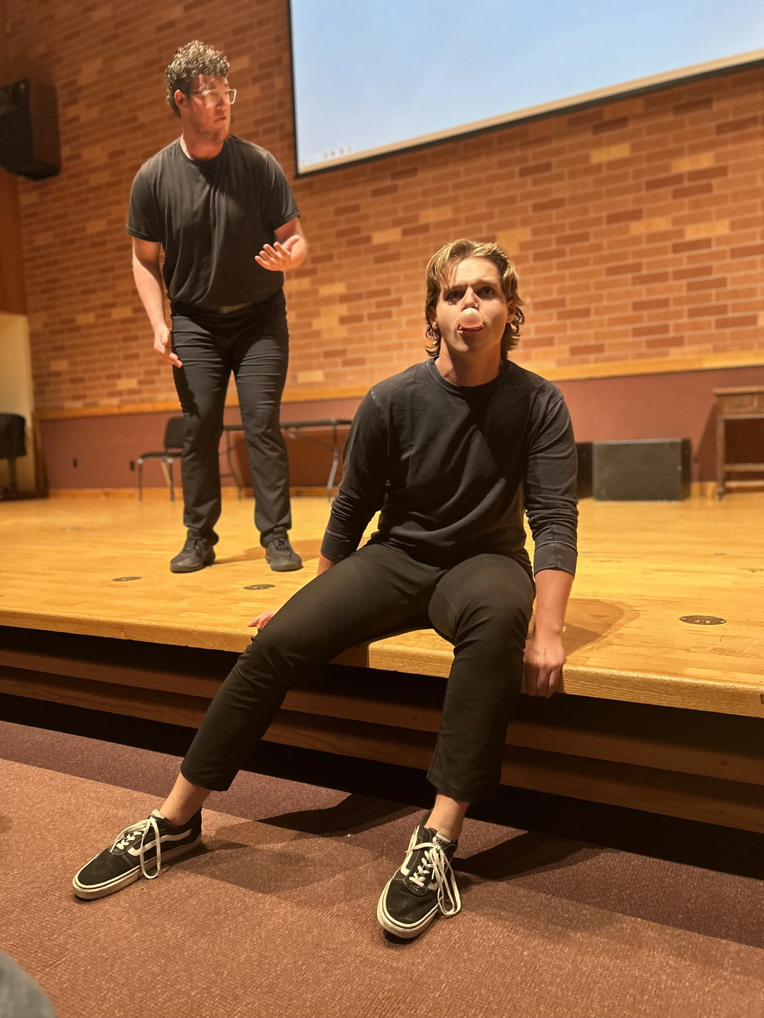 Two men are on a wooden stage in a brick-walled room. One is sitting on the edge of the stage, sticking out his tongue, while the other stands behind him, gesturing with one hand.