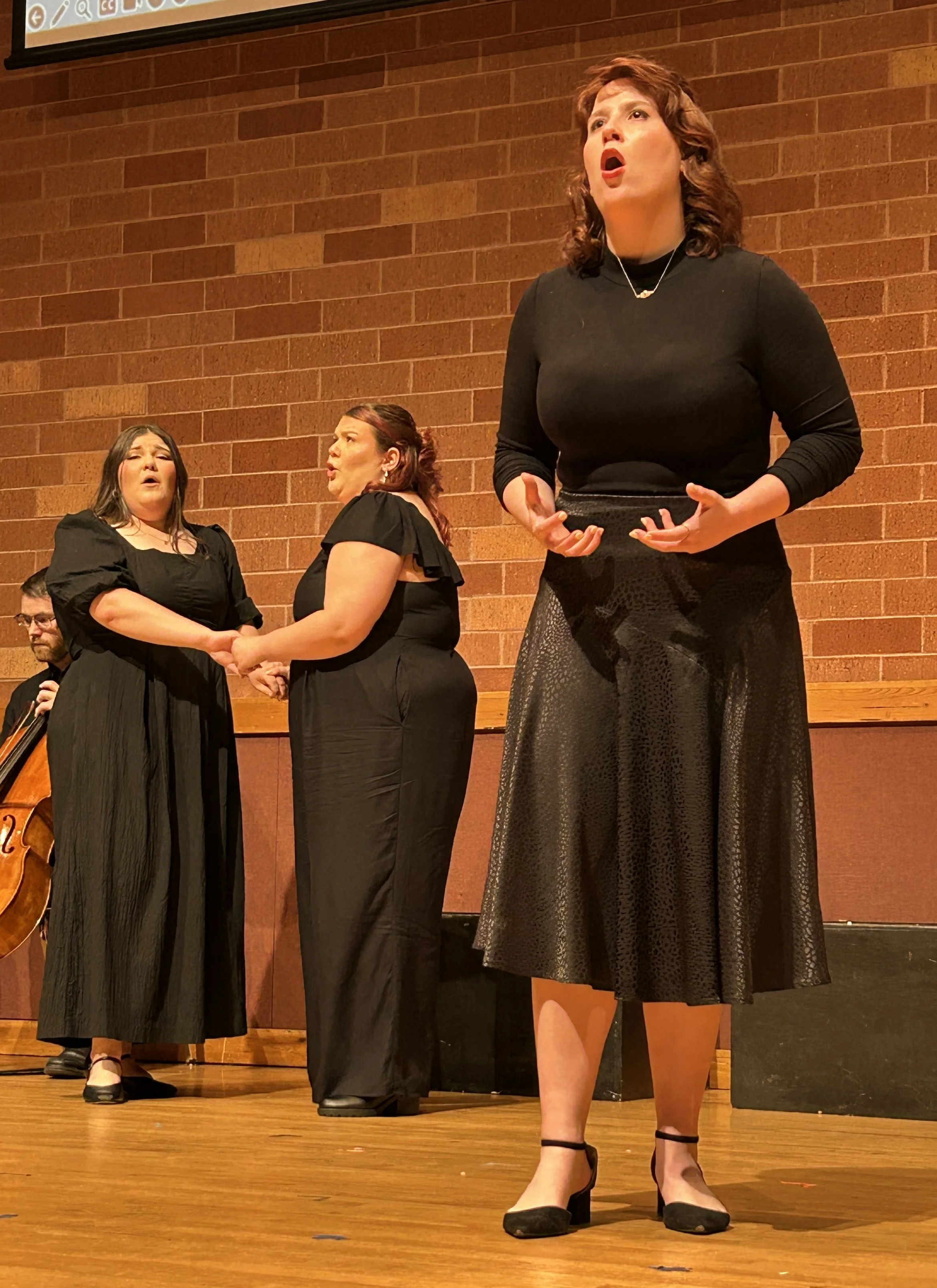 Three women singing on stage, two women holding hands in the background, with a double bass player partially visible on the left side. They are performing in front of a brick wall.