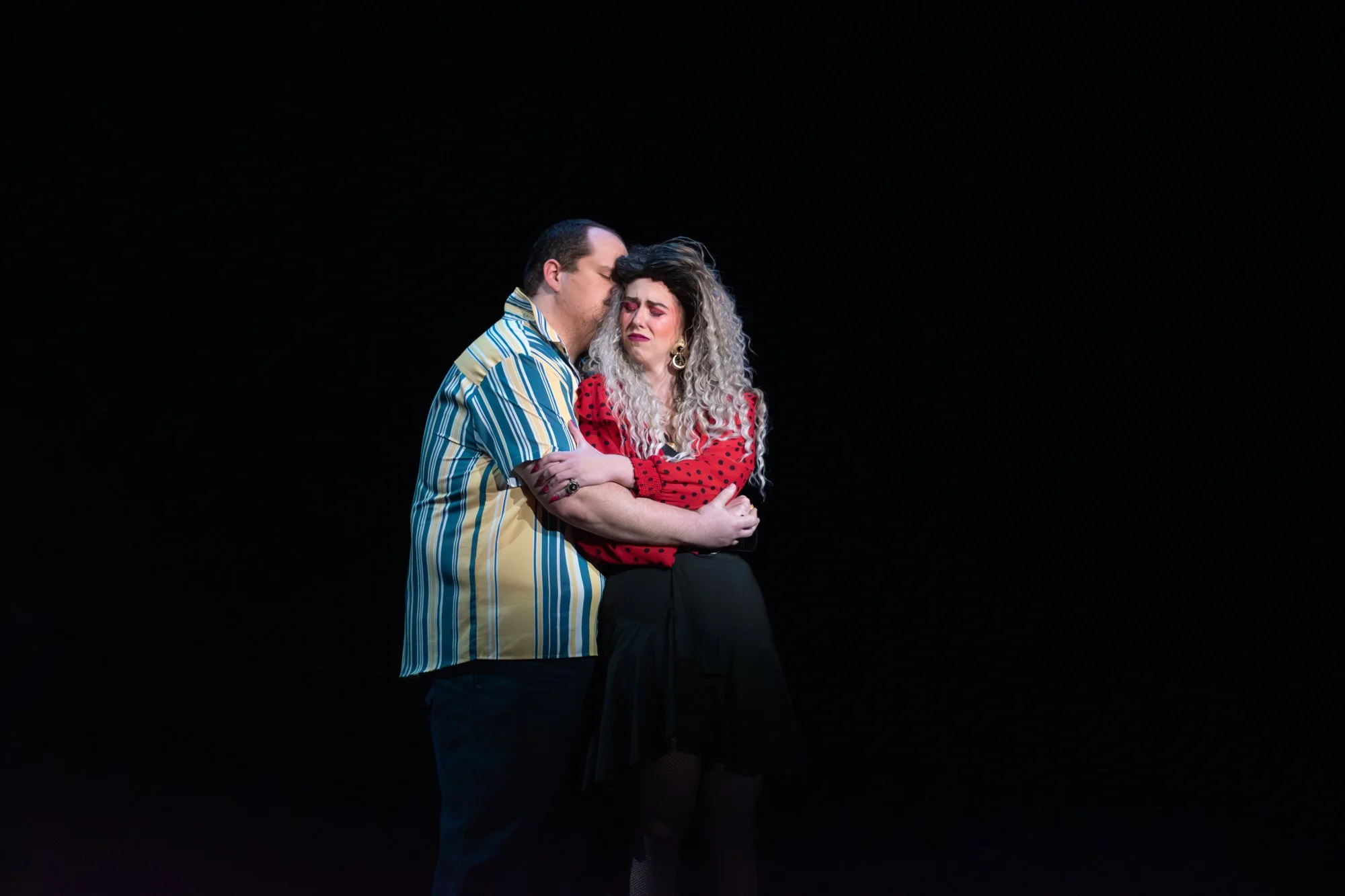Two actors standing close together on stage, embracing and showing emotional expressions, against a black background.