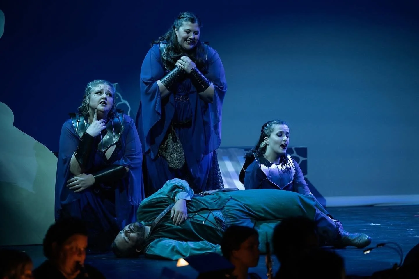 Theater scene with four women in dark blue costumes, one lying on the ground, three standing, all appearing distressed, with stage background suggestive of a storm or lightning.