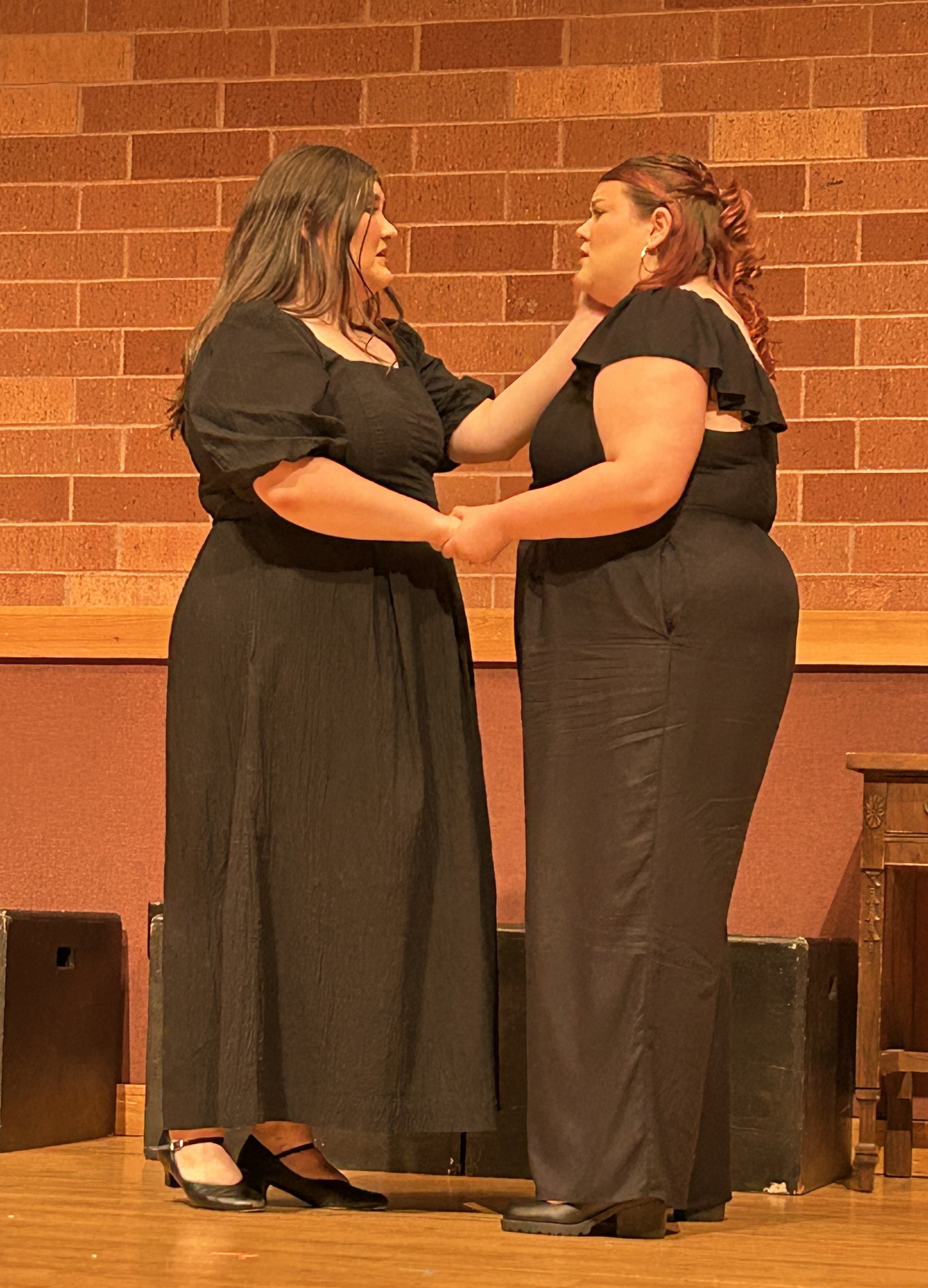 Two women on stage holding hands, one woman is touching the other's face, both women are wearing black dresses, in front of a brick wall.
