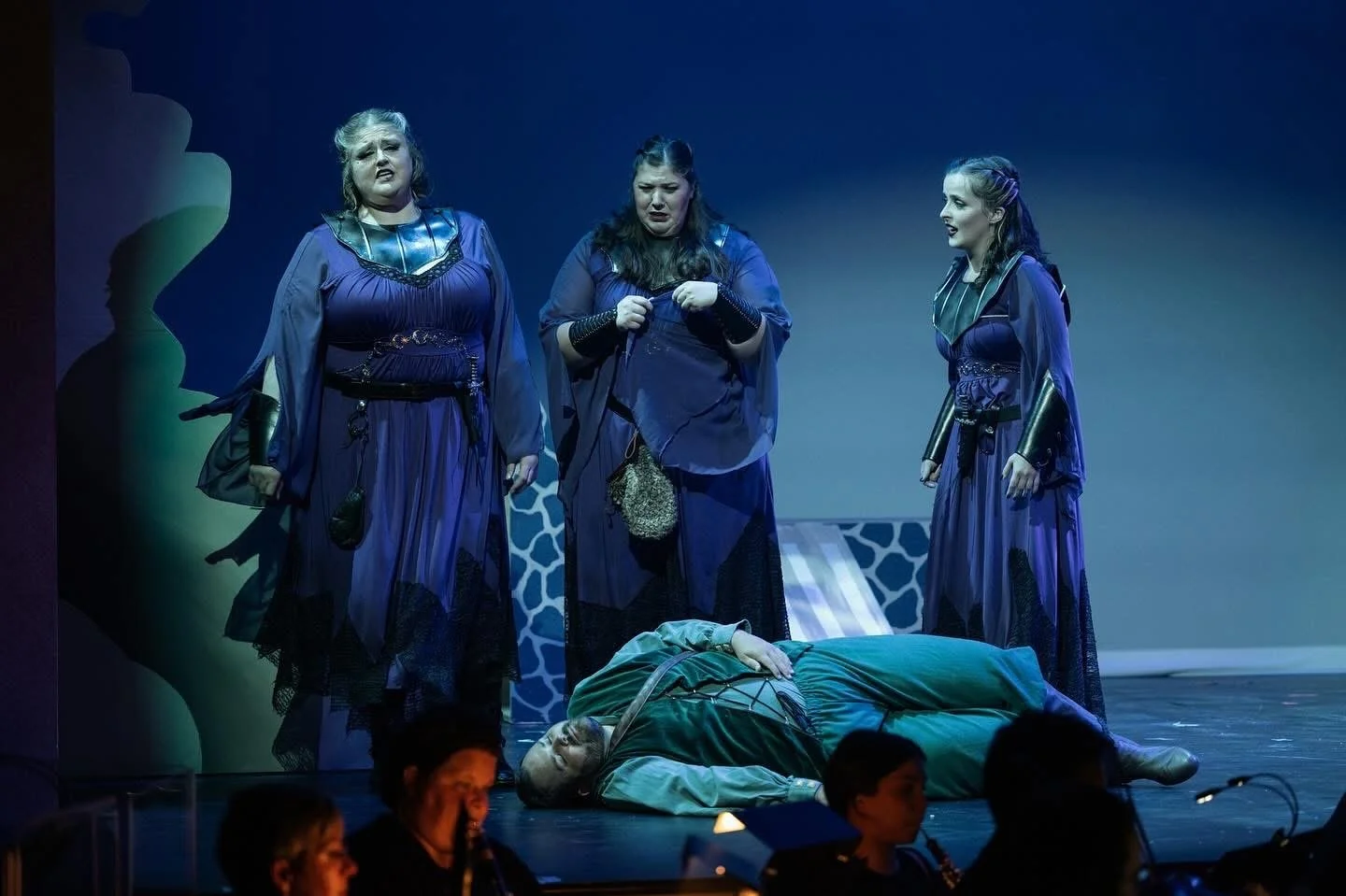 Three women in purple costumes standing on stage, one man lying on the ground in front of them, in a theatrical performance.