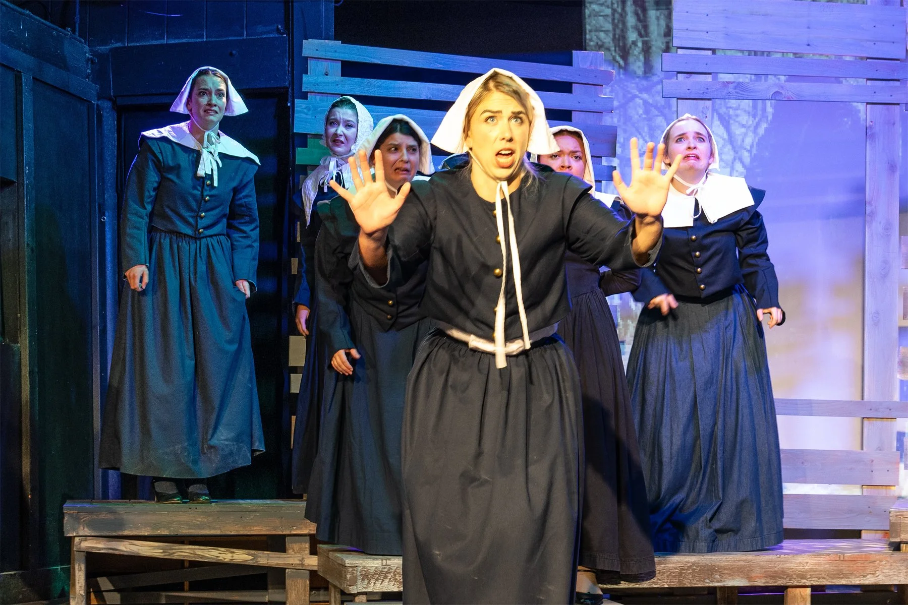Women dressed as nuns on stage during a theatrical performance, with one in front making a panicked or shocked expression and several others behind showing concern or fear.