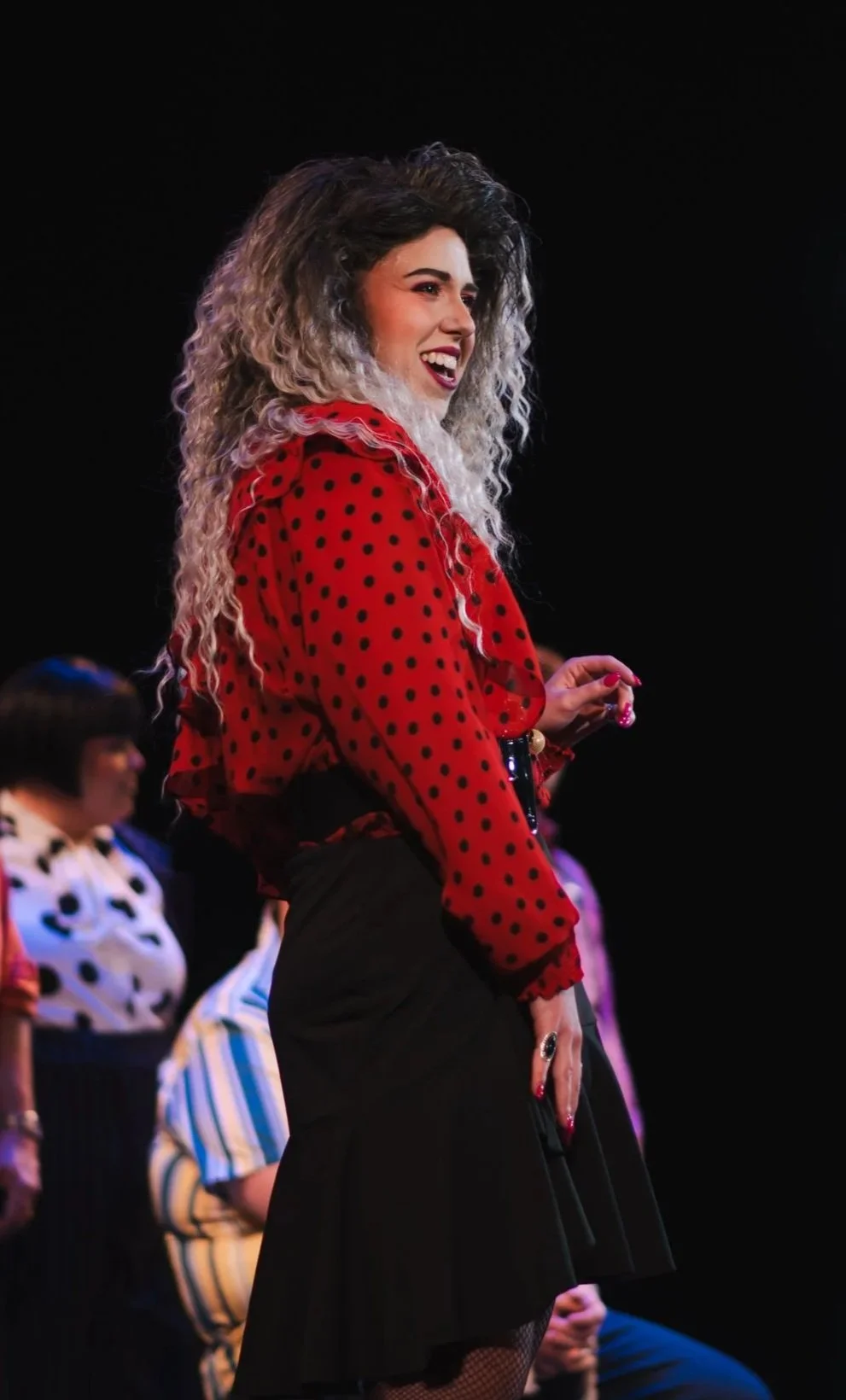 A woman with untamed, curly, ombré hair (dark at the roots, blonde at the tips) smiling on stage, wearing a red blouse with black polka dots and a black skirt.
