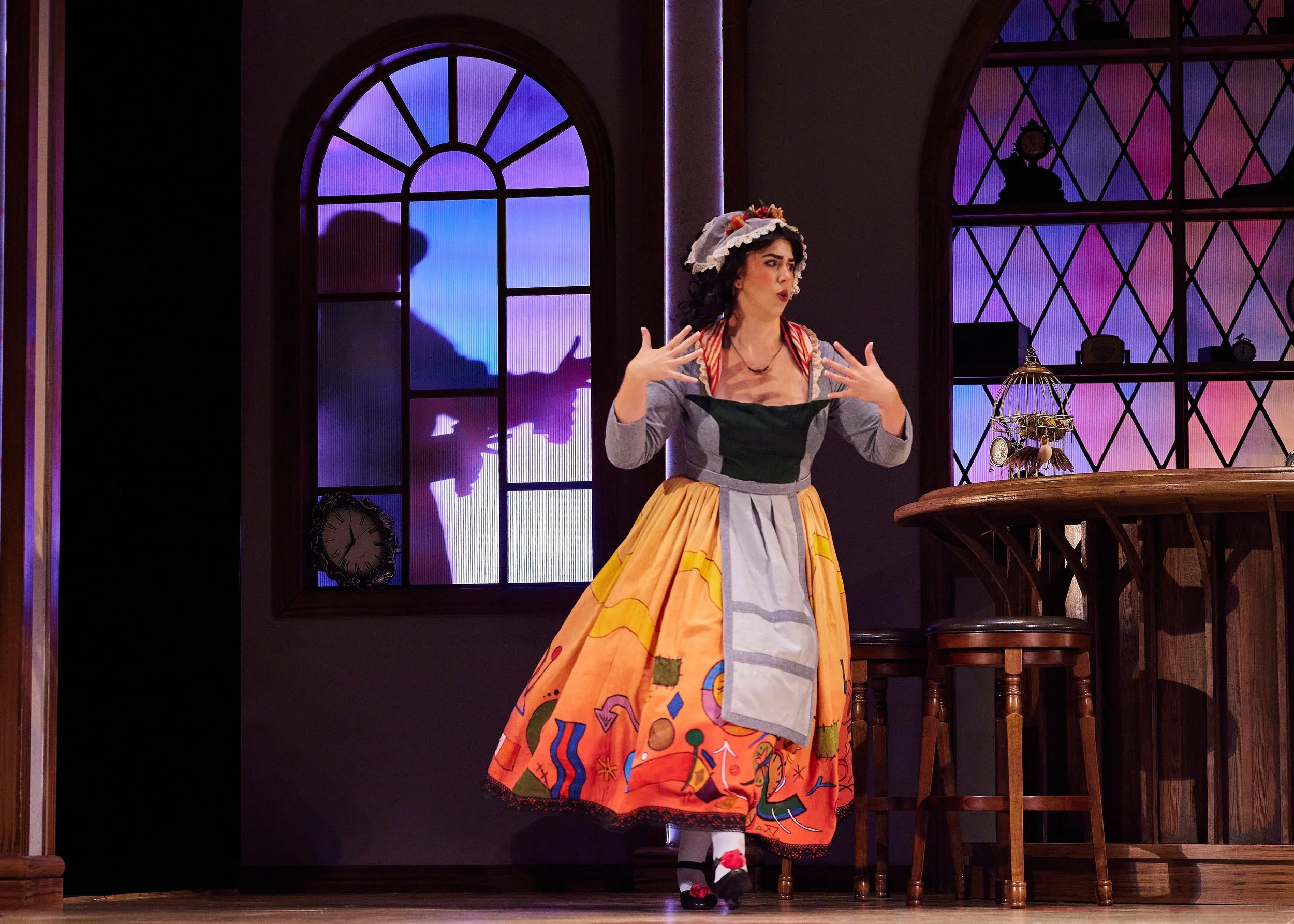 A woman dressed in a colorful costume with a bright orange skirt and a painted apron, performing on stage with a background of stained glass windows and a silhouette of a man. She is gesturing with her hands and wearing a floral hat.