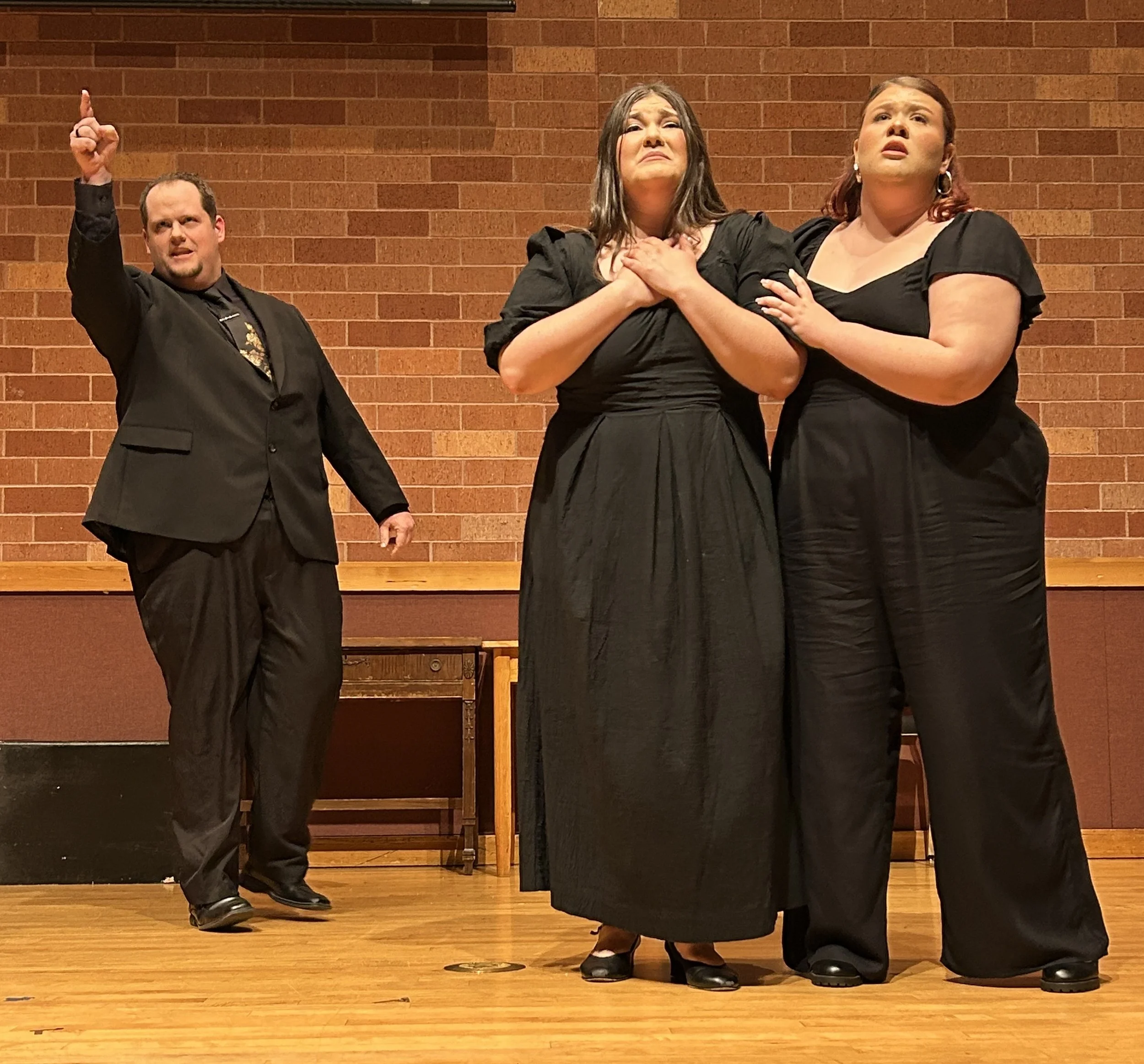 Three actors on stage, one man and two women, dressed in black formal attire. The woman in the center appears emotional, clutching her chest, as the other woman gently touches her shoulder. The man gestures with his right hand, pointing upward, looki