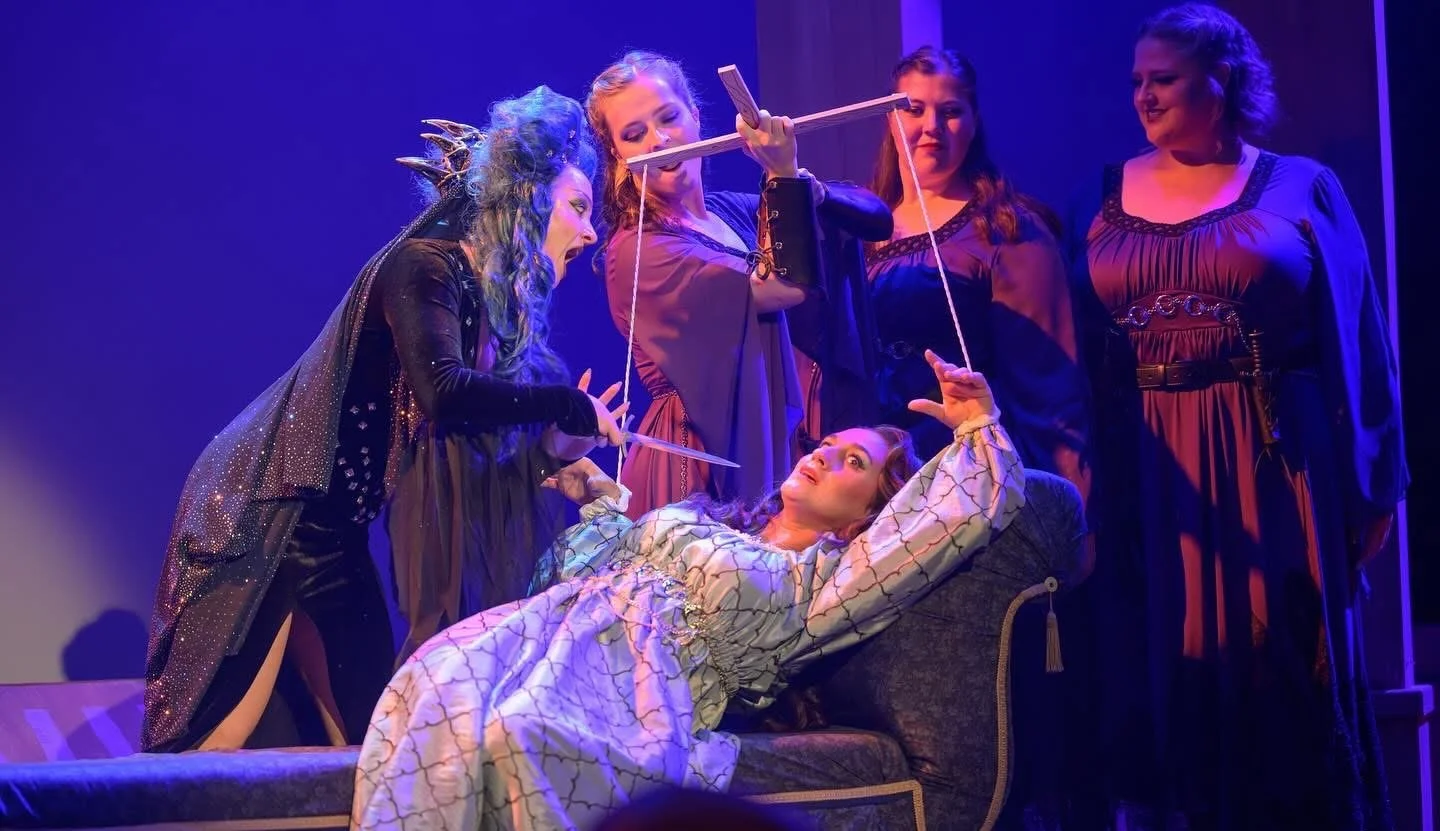 Group of women performing a theatrical scene on stage, one woman lying on a sofa and the others surrounding her, with intense expressions and dramatic costumes.