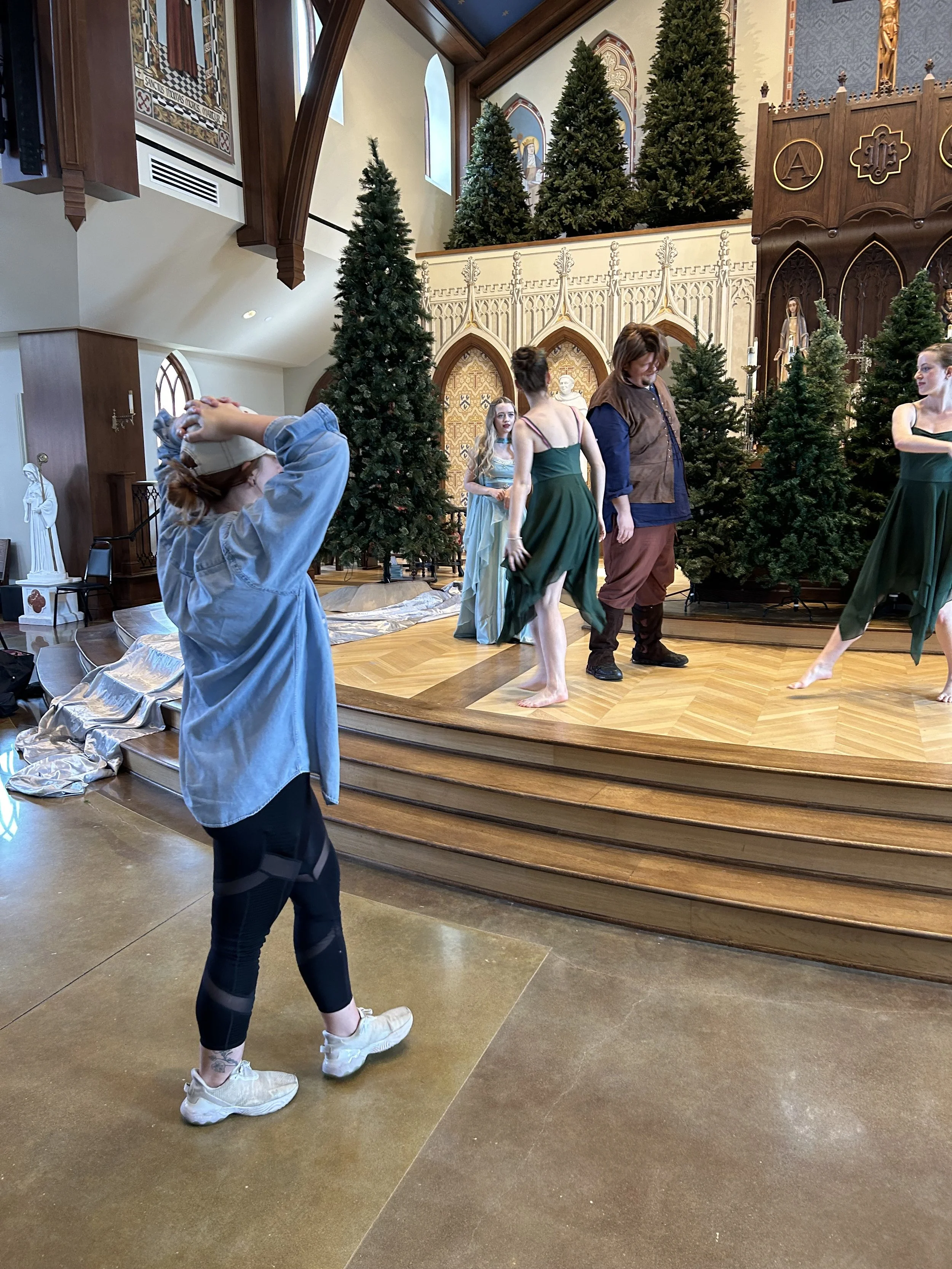 A person in casual clothing standing in front of a stage with ballet dancers, decorated with Christmas trees and religious artwork inside a church.