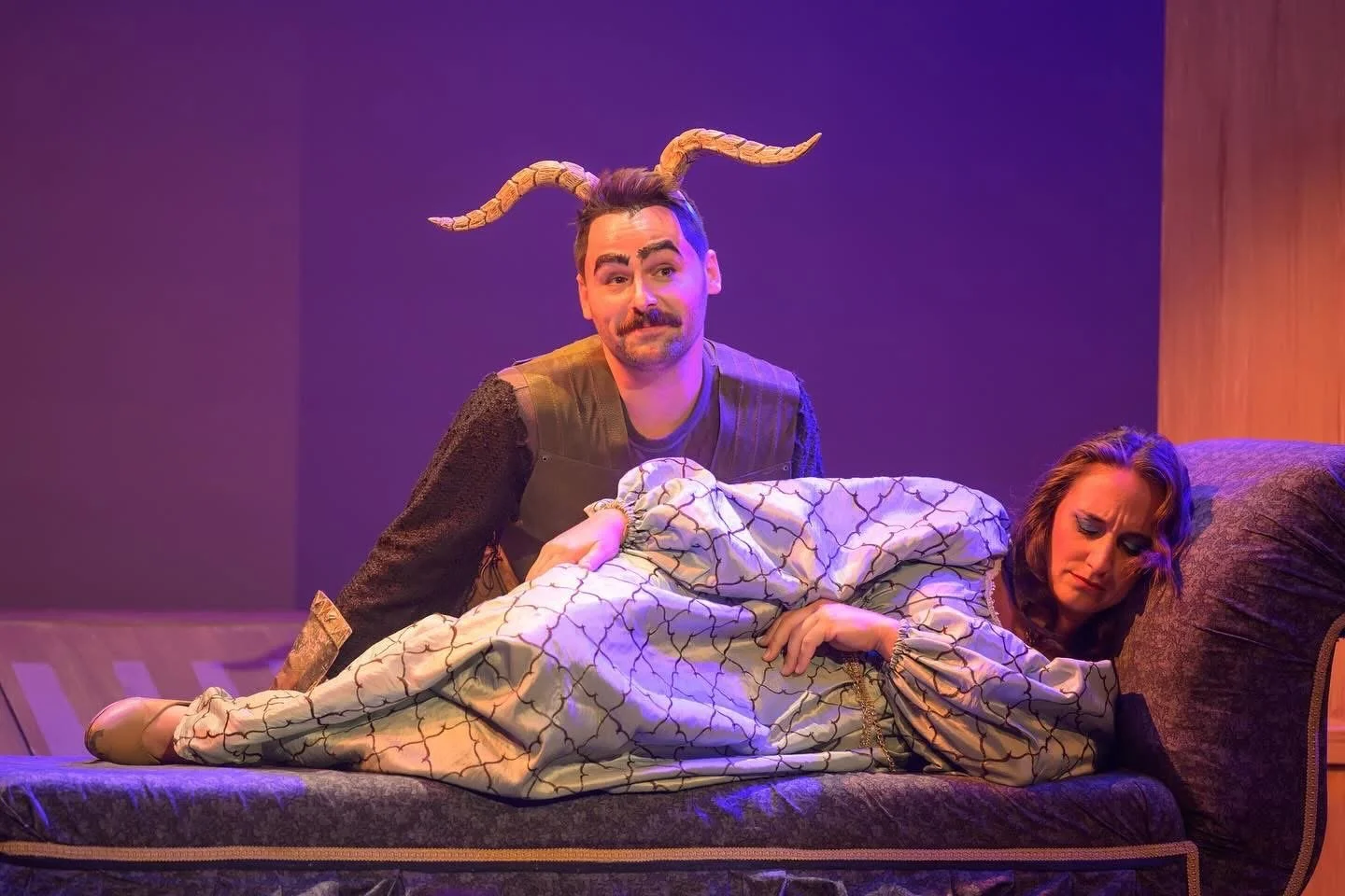A man with a mustache and bushy eyebrows wearing a costume with devil horns and a vest, sitting beside a woman with long brown hair and makeup, lying on a bed with a patterned coverlet, both on a theater stage with purple and orange lighting.