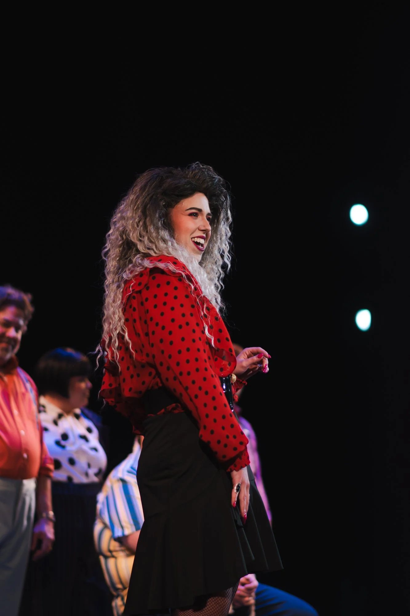 A woman with curly hair in a red polka dot blouse and black skirt standing on stage, smiling and looking to the side, with a group of people behind her.