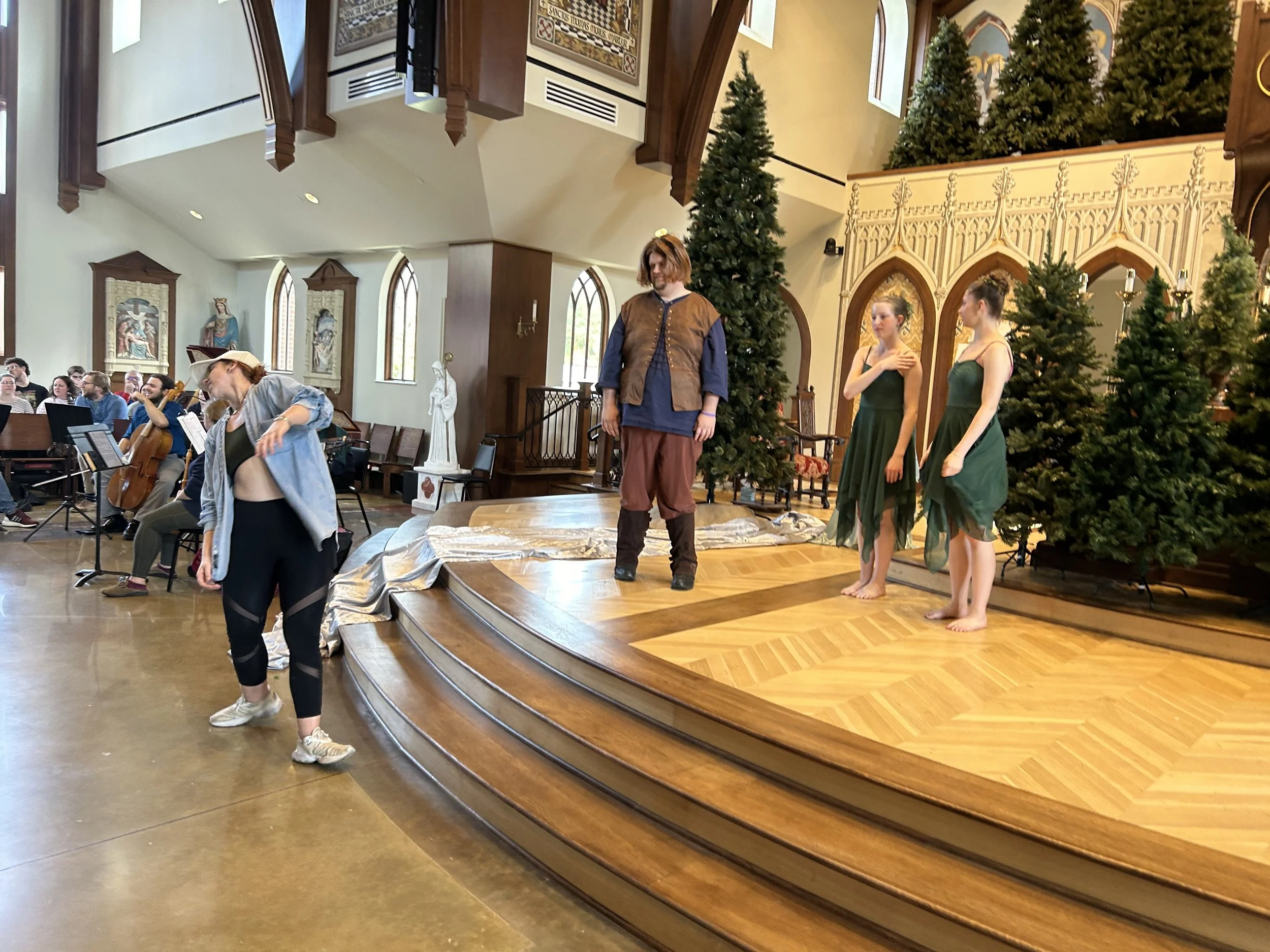 Rehearsal or performance taking place in a church with actors on stage and musicians in the background, decorated with Christmas trees.