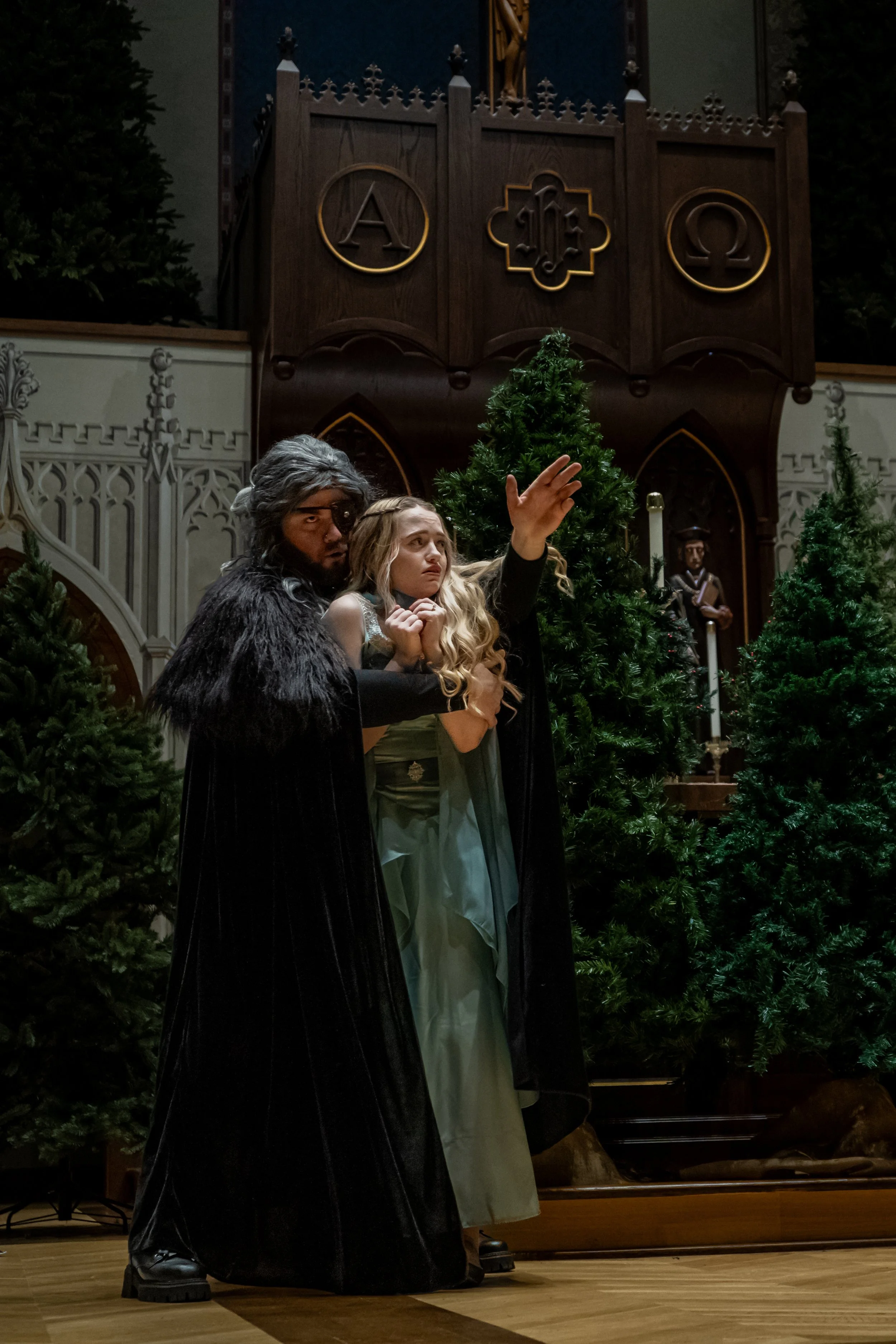 Two actors in costume perform on stage inside a church, with Christmas trees and religious symbols in the background.
