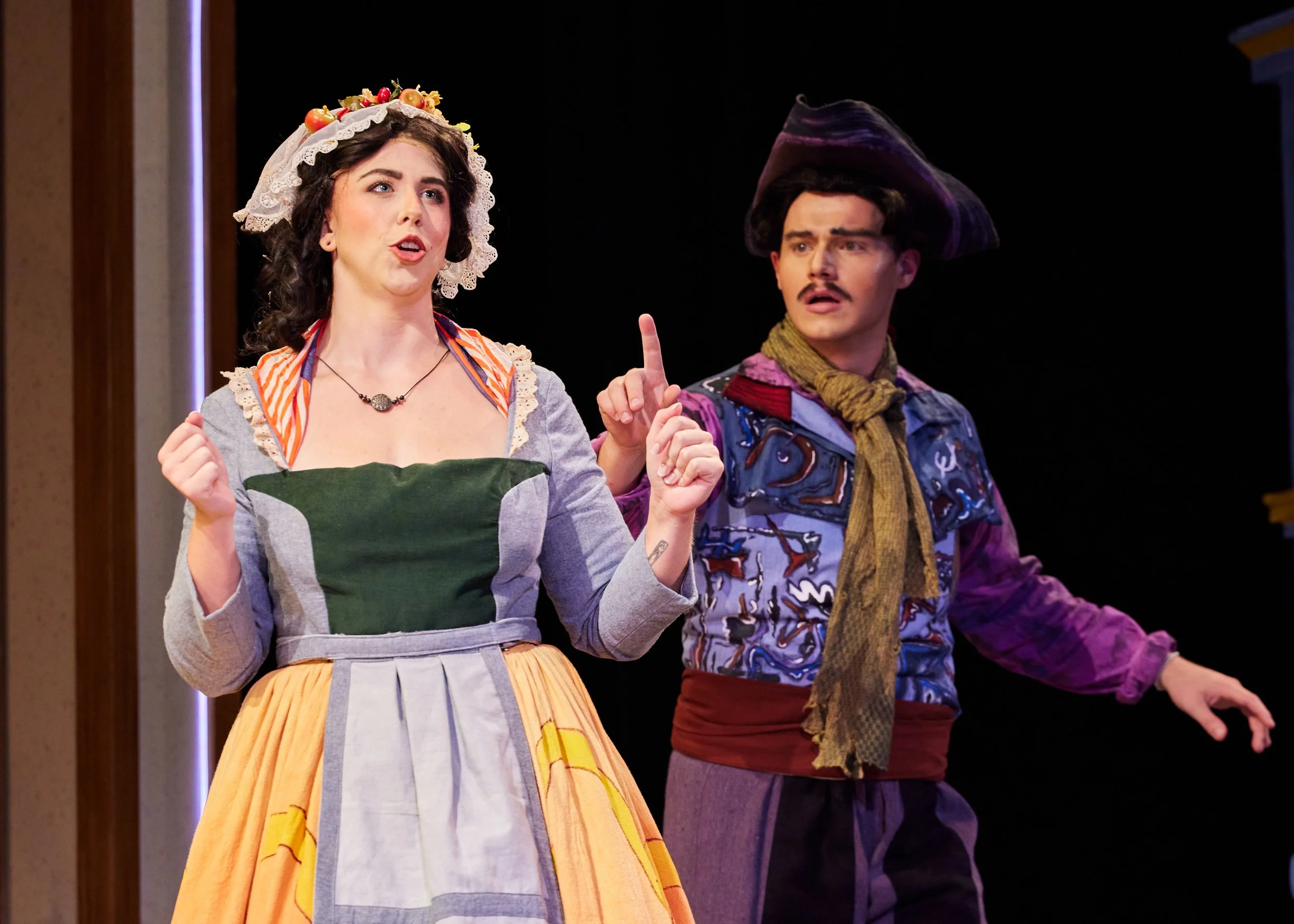 Two actors in costume perform on stage, with one woman wearing a dress with an apron and a bonnet decorated with fruit, and a man dressed as a cowboy with a large hat, scarf, and colorful shirt.