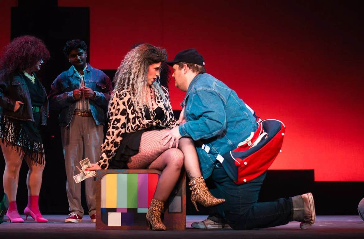 The scene is from a theatrical play with five actors on stage. The main focus is a woman with curly blonde hair, leopard print top, fishnet stockings, and leopard print boots, sitting on a colorful block. She appears to be serenaded by a man kneeling