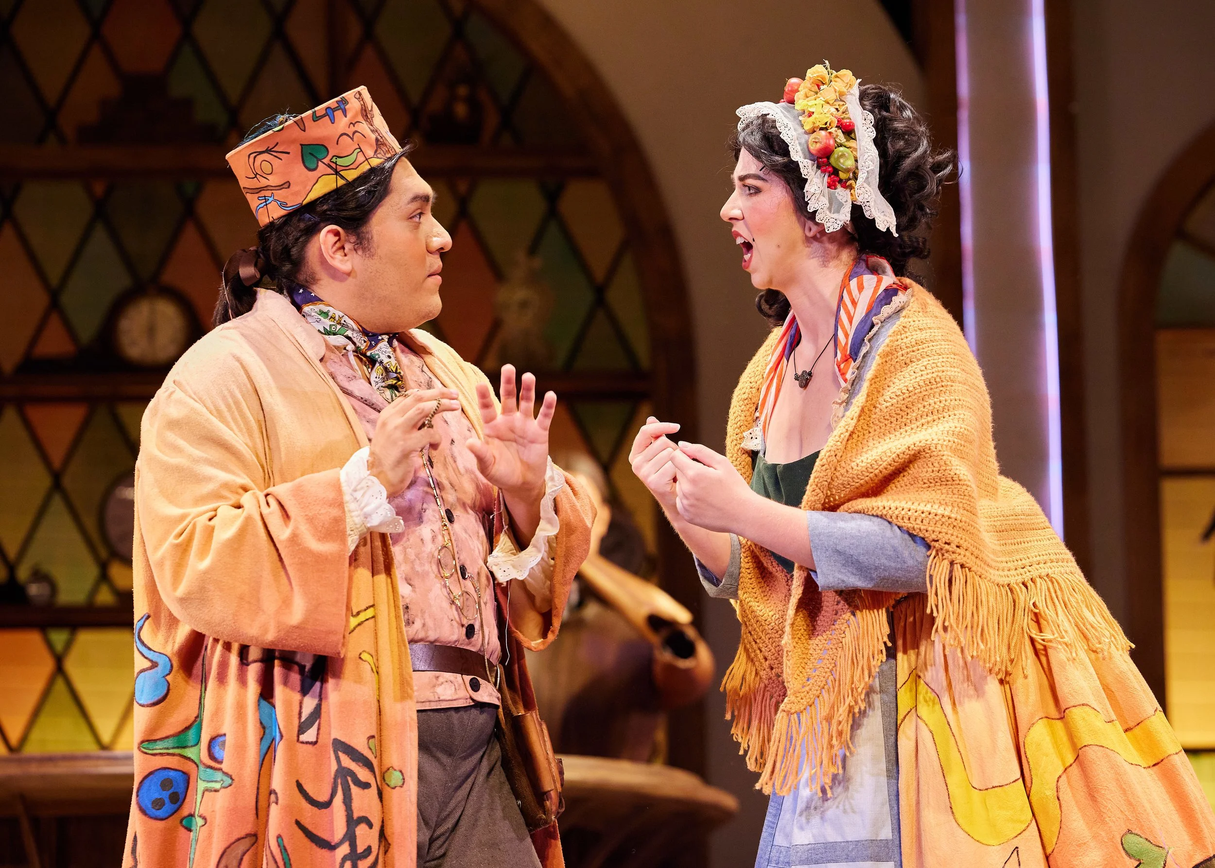 Two actors performing in a theatrical play, dressed in colorful, patterned costumes with expressive facial emotions, indoors with a decorative background.