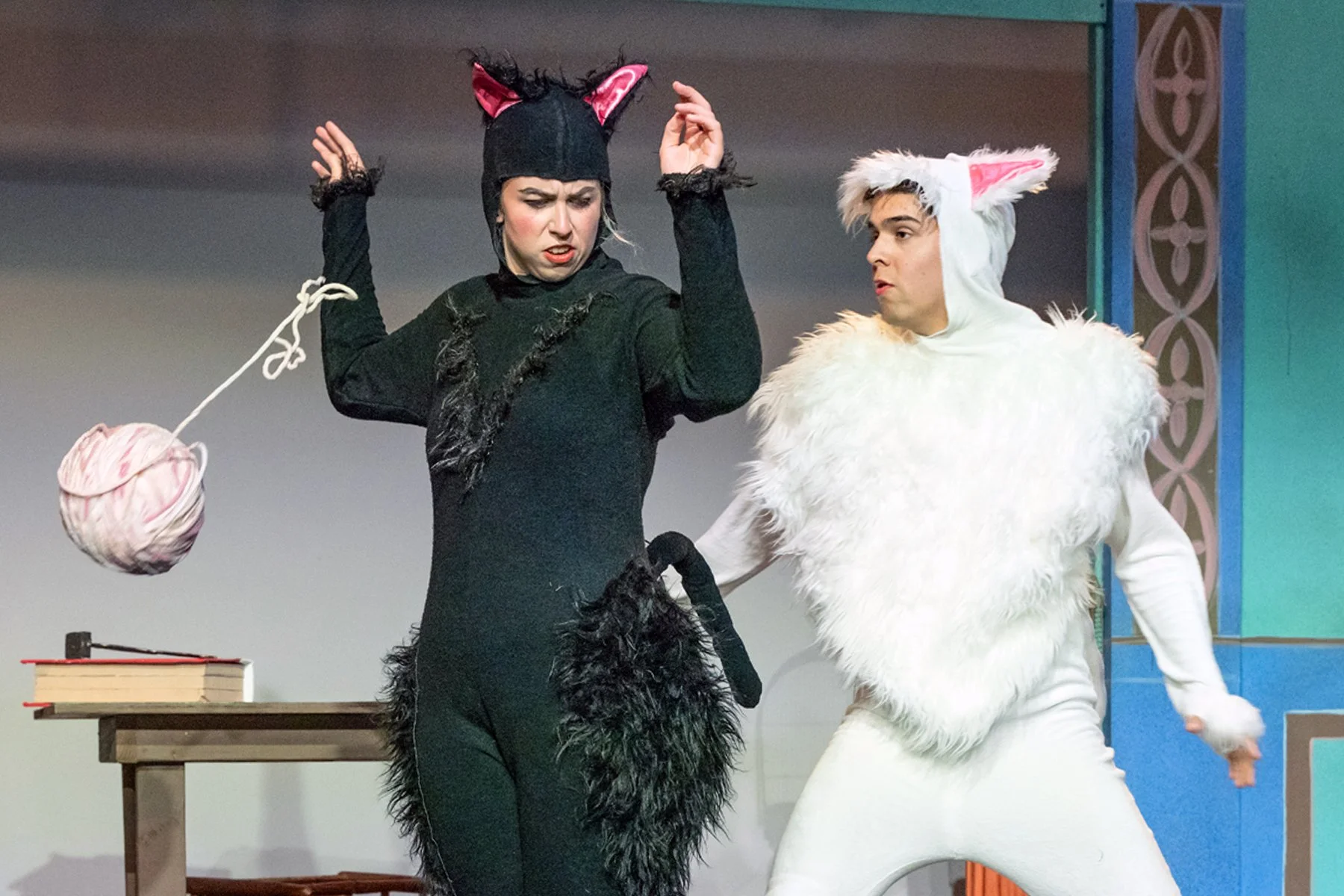 Two actors in a theatrical performance wearing cat costumes, one in black with pink ears and a tail, the other in white fur with pink ears, on stage with a small table and books in the background.