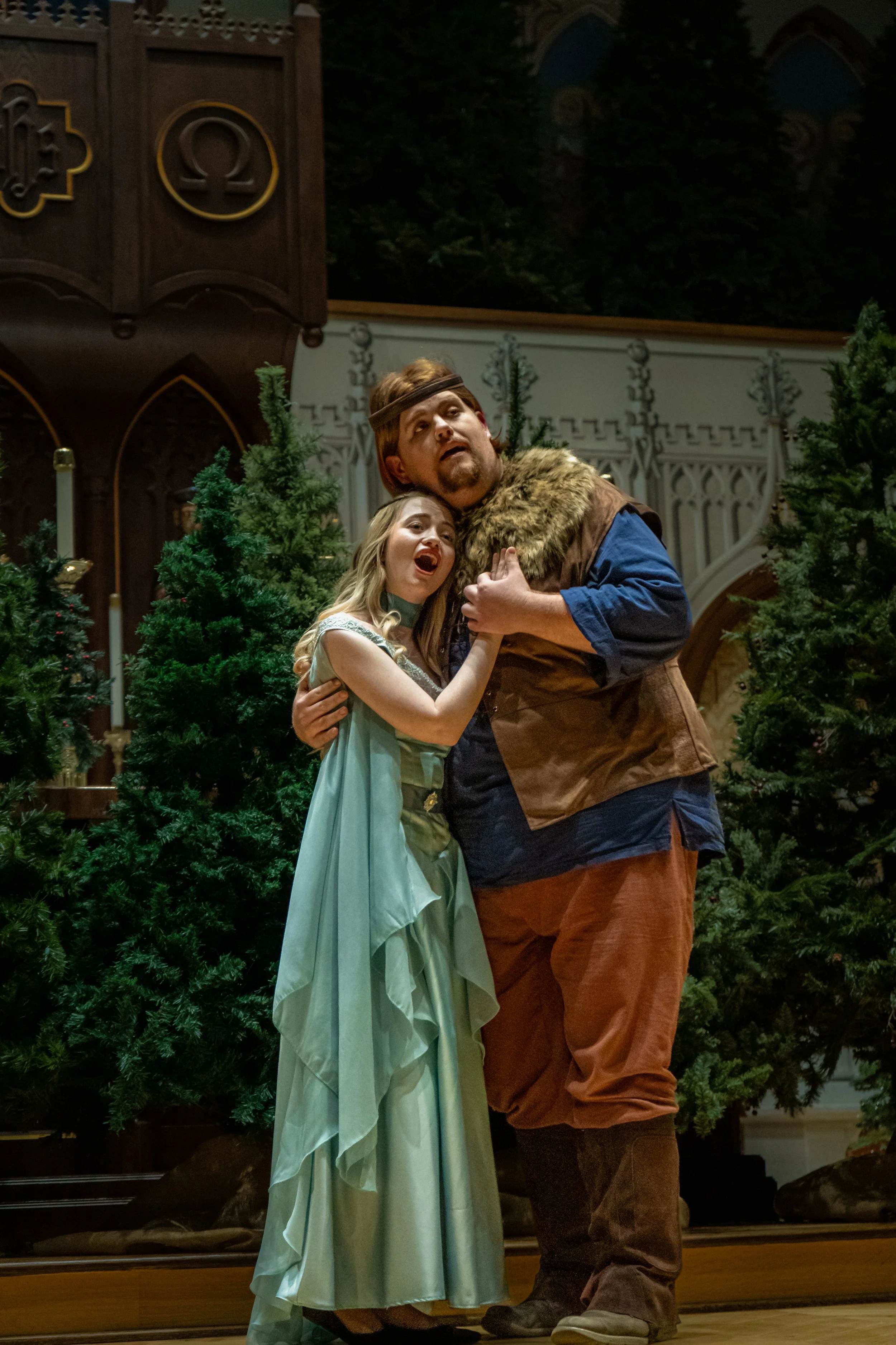A theatrical scene with a girl in a light green princess dress and a man in medieval costume hugging each other, surrounded by Christmas trees and set decorations.