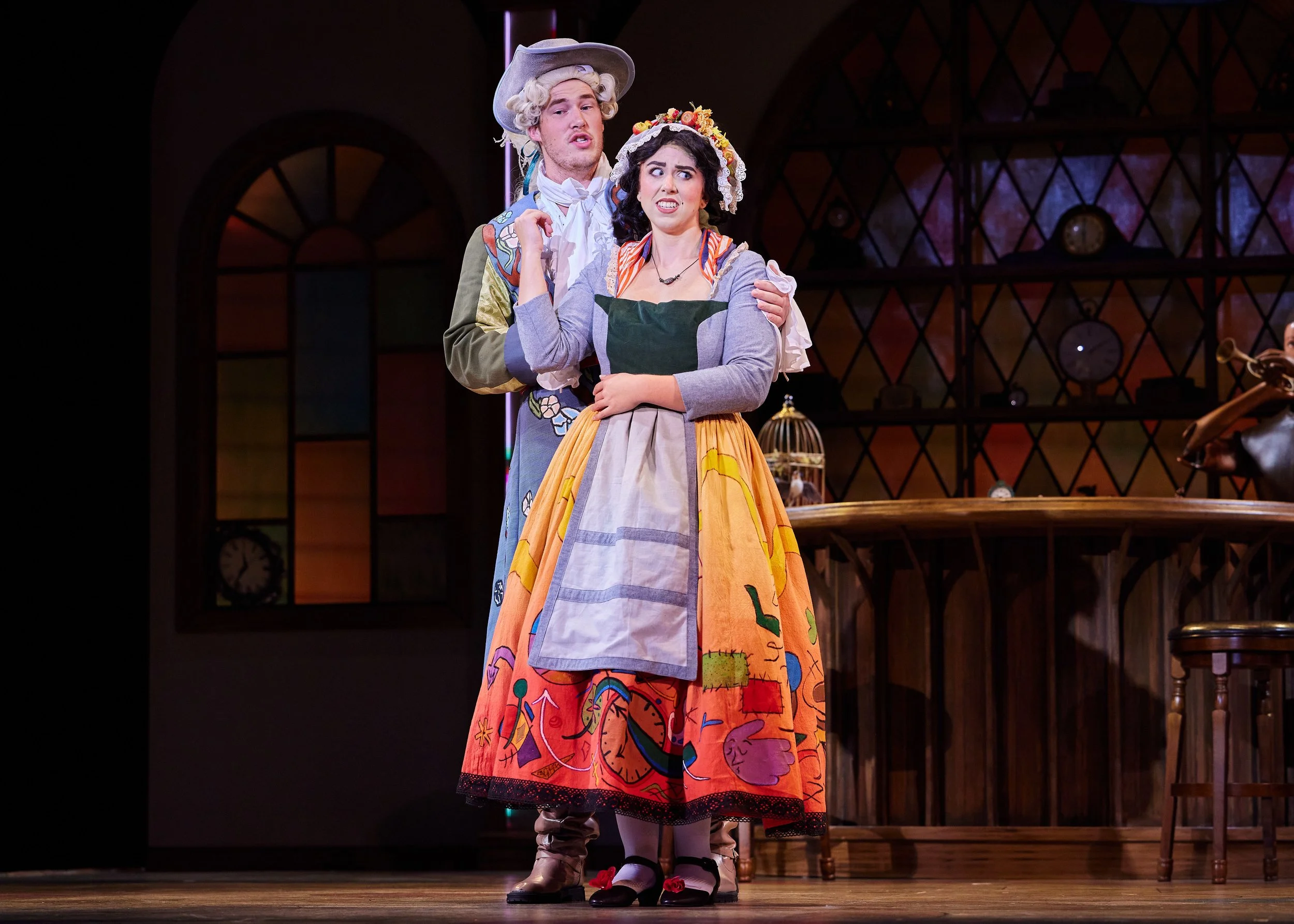 Two actors on stage dressed in colorful costumes, with one in a floral skirt and apron and the other in a suit with a large hat, performing in a theatrical play.
