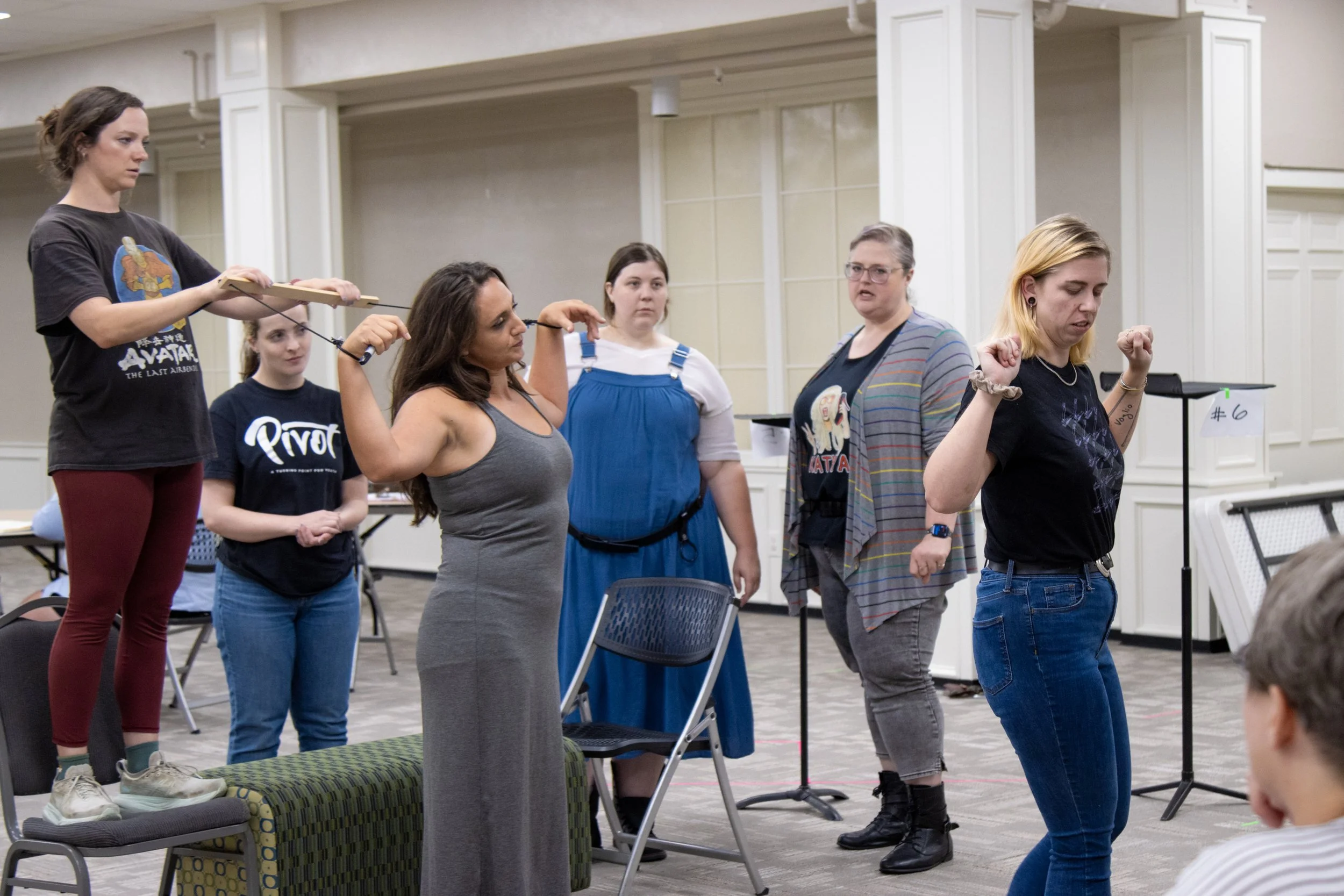 Women participating in a theater rehearsal or acting class, with some standing and some sitting, appear focused and engaged.