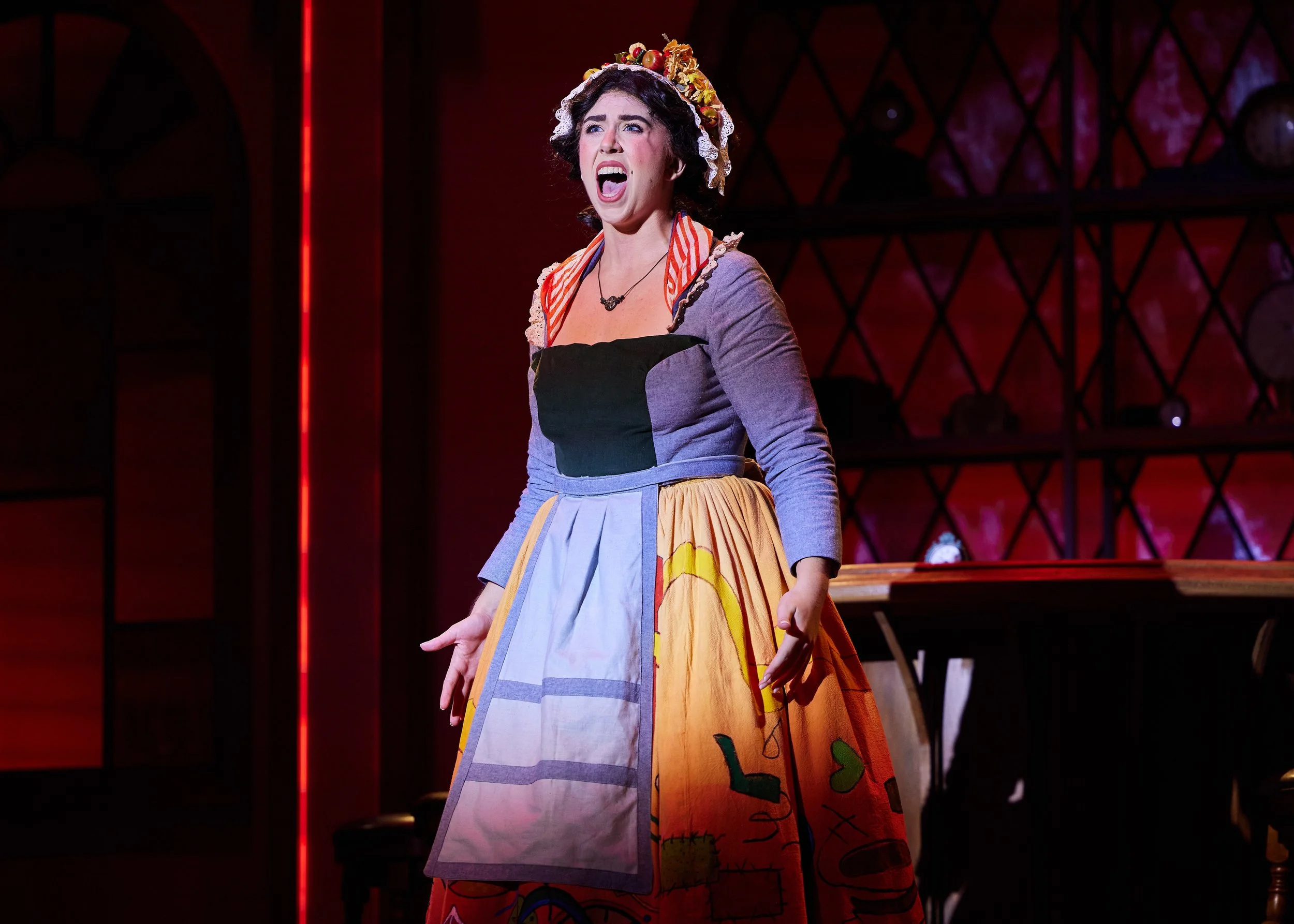 Stage actress in colorful costume, singing passionately, with a dramatic red and wooden background.