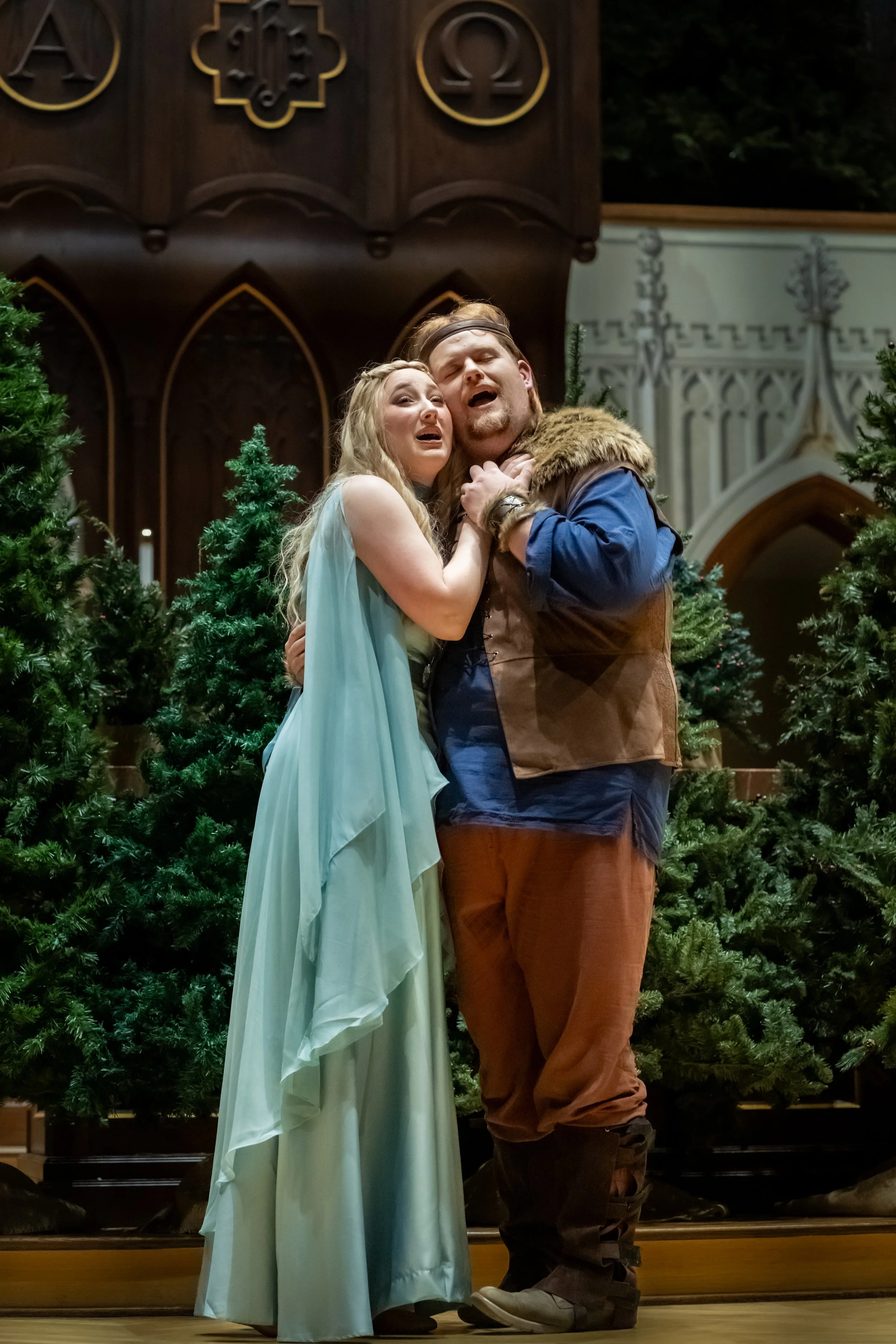 Two actors on stage dressed in costume, with Christmas trees in the background, performing a dramatic scene with emotional expressions.