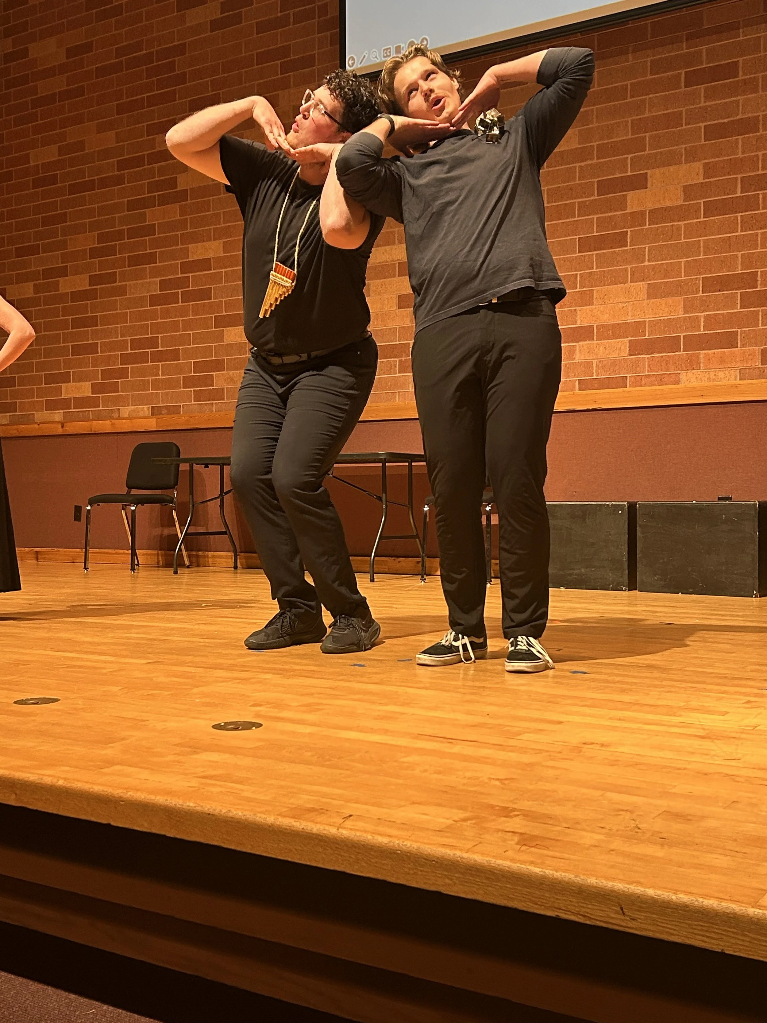 Two men standing on a wooden stage, making expressive gestures with their hands near their faces, while performing or acting. One wears glasses and a necklace, the other has a beard and is wearing sneakers.