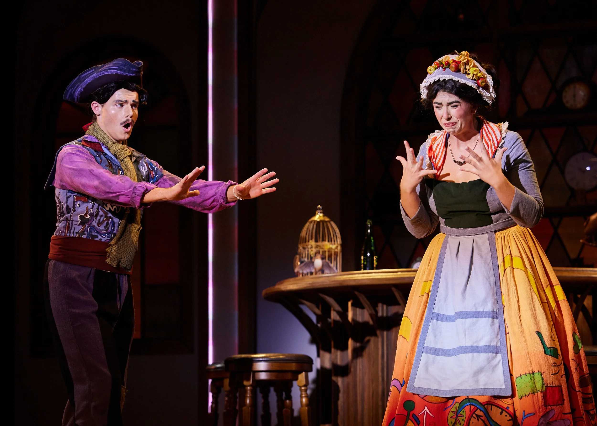 Two actors perform on stage, the man on the left gestures with open hands, wearing a purple hat and Victorian-style clothing, and the woman on the right, dressed in a colorful skirt and a white apron, appears distressed with her hands near her face.