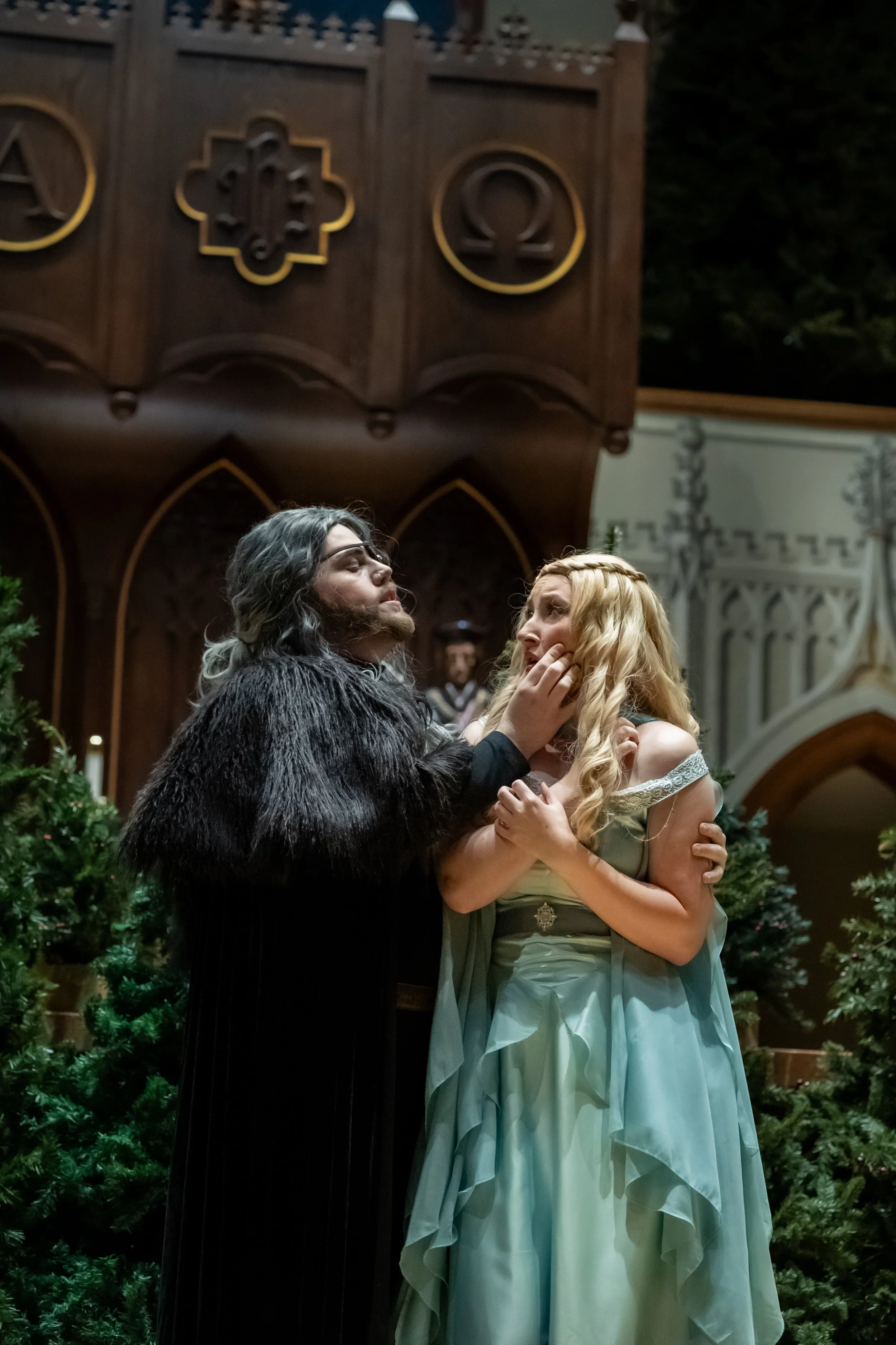 A scene from a fantasy-themed wedding or event with two characters in costume, one with dark hair in a black fur-like cloak and the other with long blonde hair in a flowing gown, standing close together in a decorated setting with greenery and Gothic