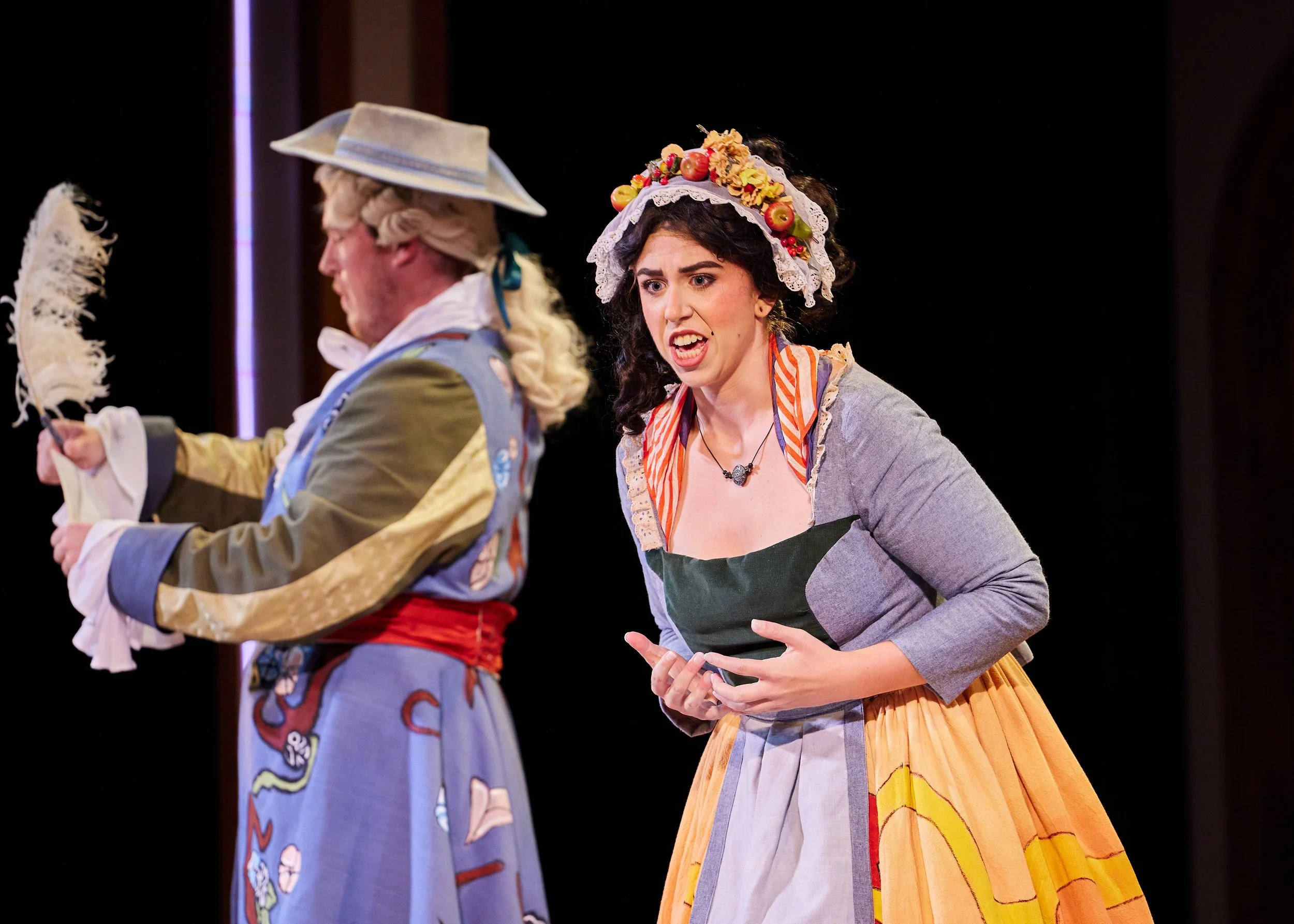 Two performers in traditional European costumes on stage; one man in a colorful, embroidered outfit with a large hat and feather, and a woman with a floral headpiece and a multicolored skirt, both appearing to act out a comedic scene.