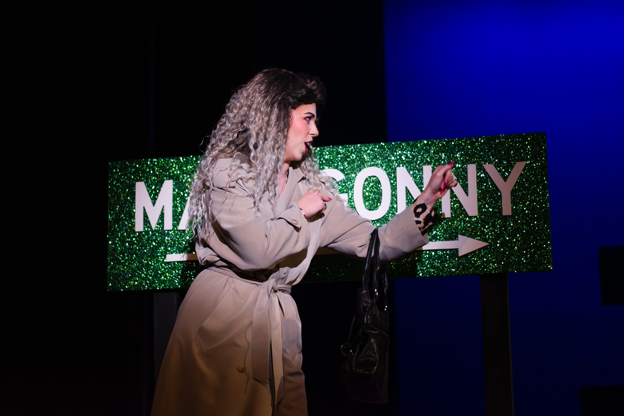 A woman with curly, blonde hair in a beige trench coat points at a green sparkly directional sign that reads 'MAIN GALLERY' with an arrow pointing to the right, on a dark background.