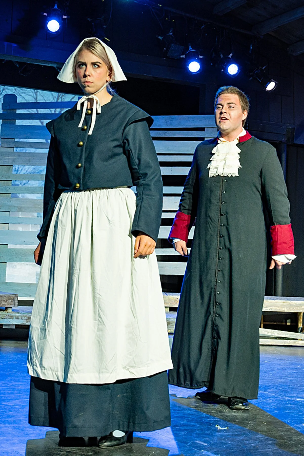 Two actors in period costumes on a stage with wooden backdrop and stage lighting. The female actor in the foreground wears a dark dress with a white apron and bonnet, looking upset. The male actor in the background wears a dark coat with red cuffs an