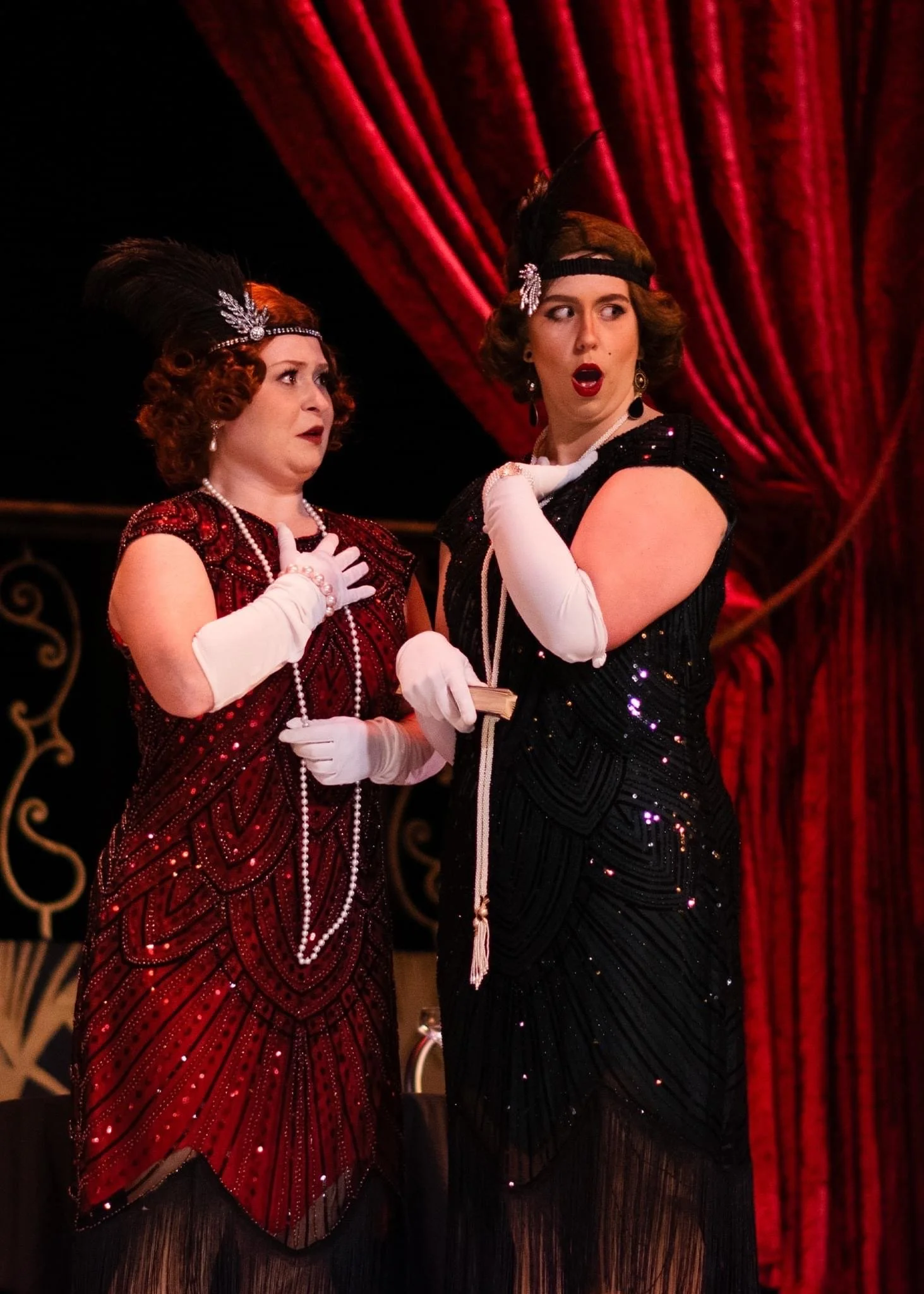Two women dressed as flappers with vintage 1920s style clothing, feather headbands, and long pearl necklaces, standing on stage with red velvet curtains in the background, displaying surprised or shocked expressions.