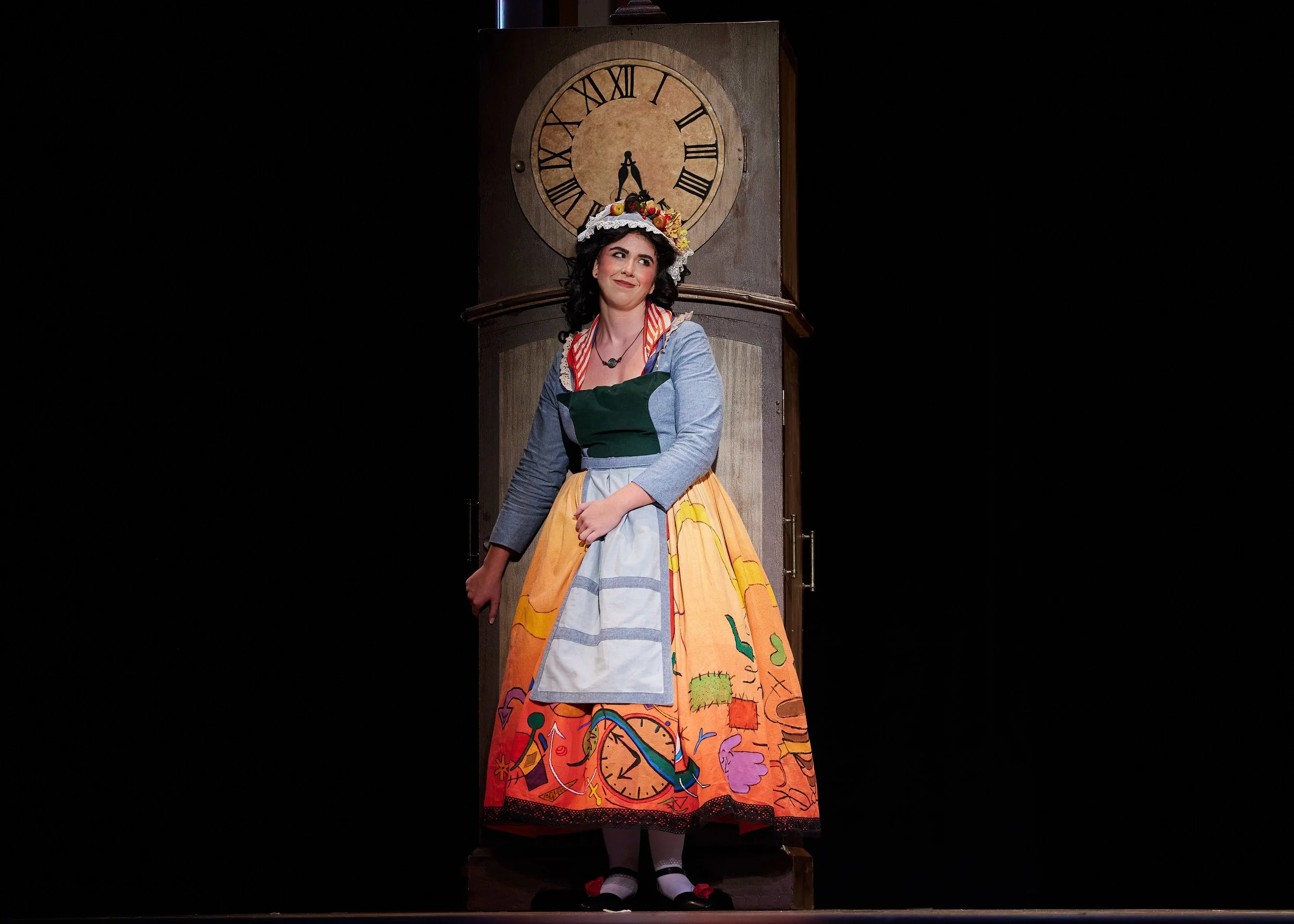 Woman in colorful costume standing in front of a large clock on stage.