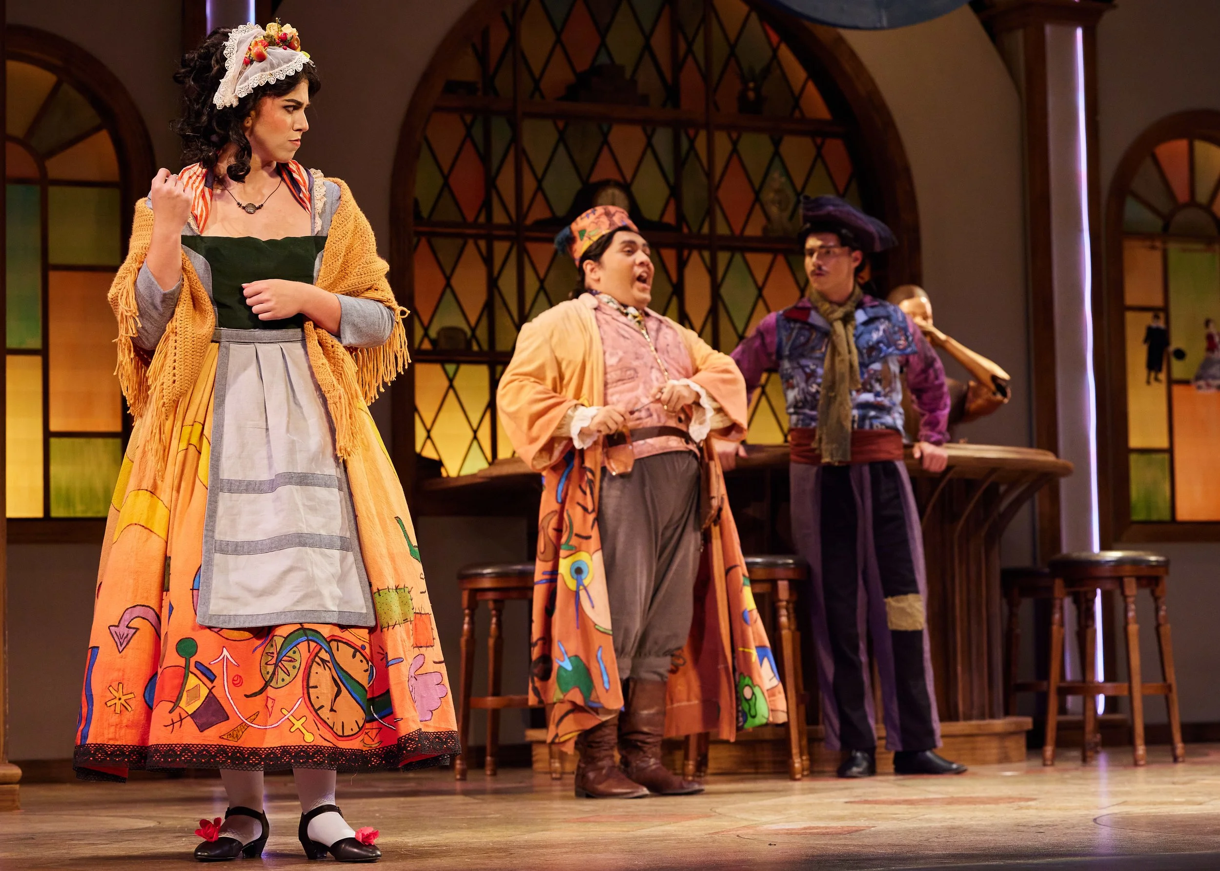Actors in colorful costumes performing on stage, with a woman in a decorated dress and two men in pirate-like outfits, in front of a set with stained glass window designs.