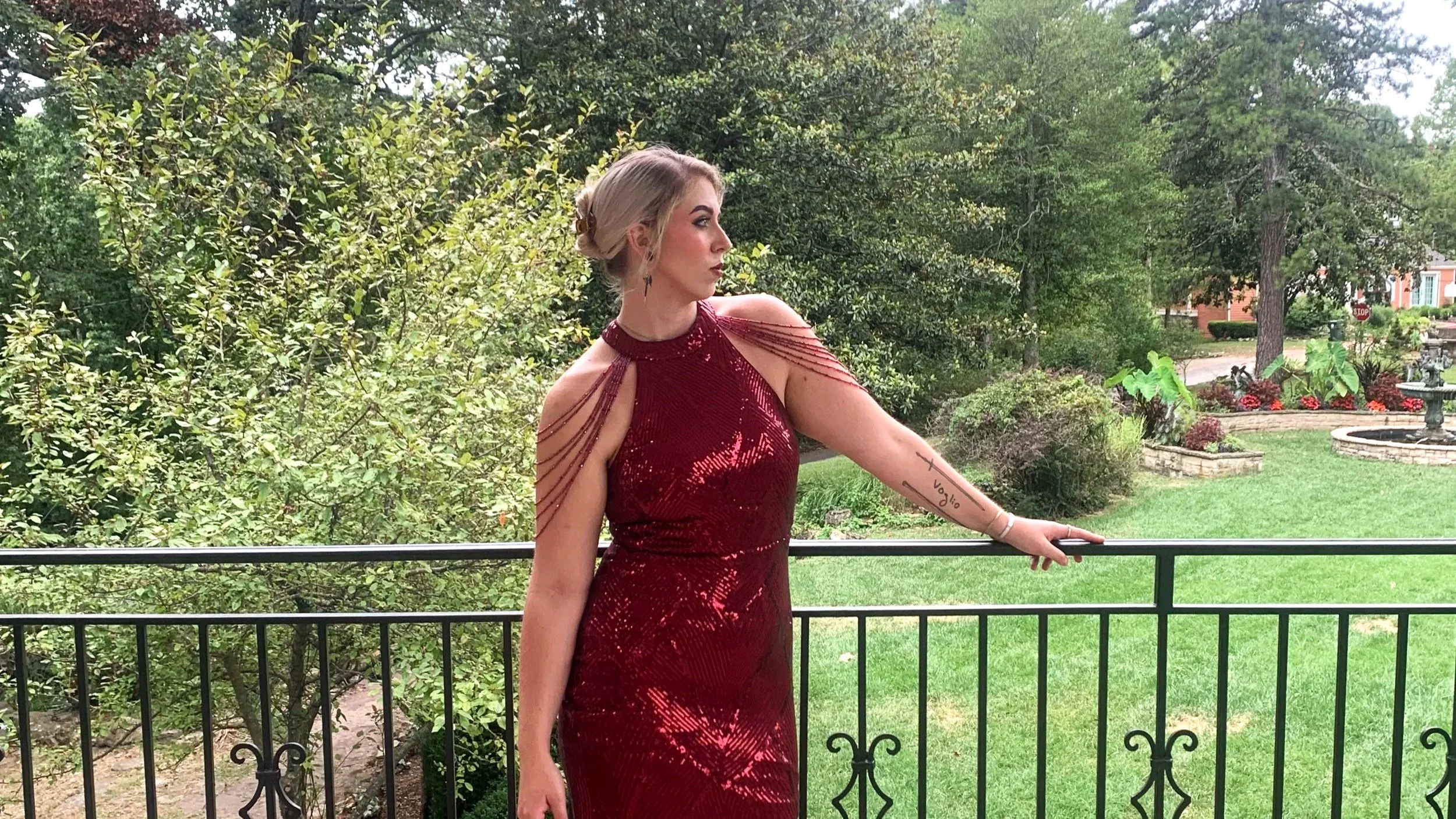 A woman in a shiny red dress with open shoulders and shoulder straps stands outside by a black metal railing. She has blonde hair styled in an updo and is wearing makeup. She has a tattoo on her left forearm. The background includes a lush green garden with trees, shrubs, a fountain, and a brick house with a stop sign are visible in the distance.