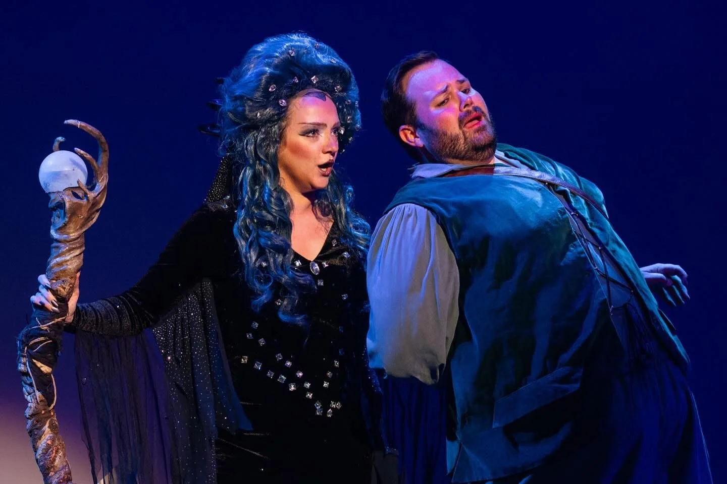 Two actors performing on stage. The woman has long, curly hair and is dressed as a witch, holding a staff. The man is wearing medieval-style clothing and is leaning back, appearing to be in a dramatic moment.