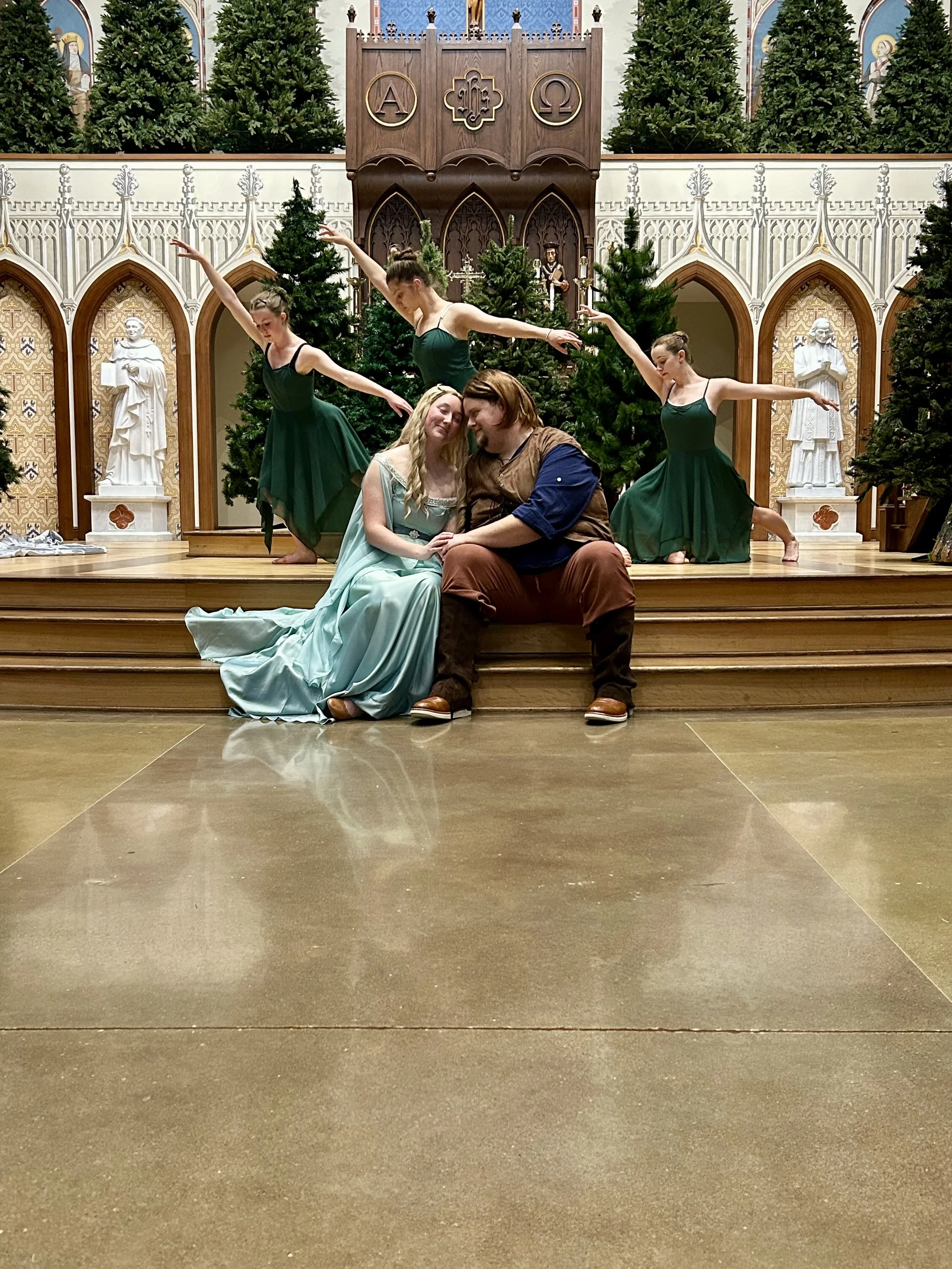 The image shows a stage with five ballet dancers practicing or performing in front of Christmas trees and religious statues in a church-like setting. Two people, a woman in a light green dress and a man in medieval-style clothing, are seated on the s