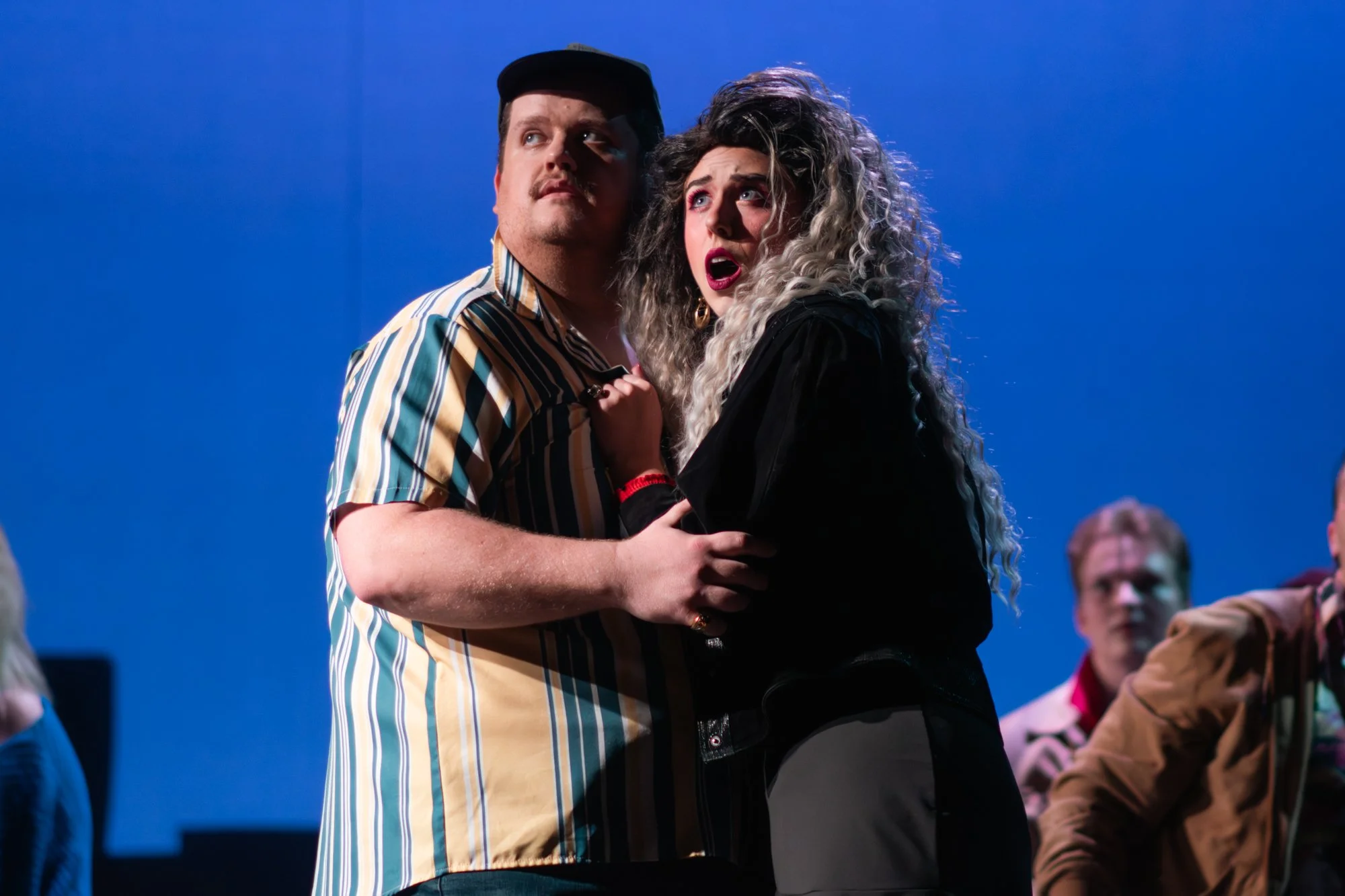 Two people, a man and a woman, are holding each other with concerned or surprised expressions, against a plain blue background.