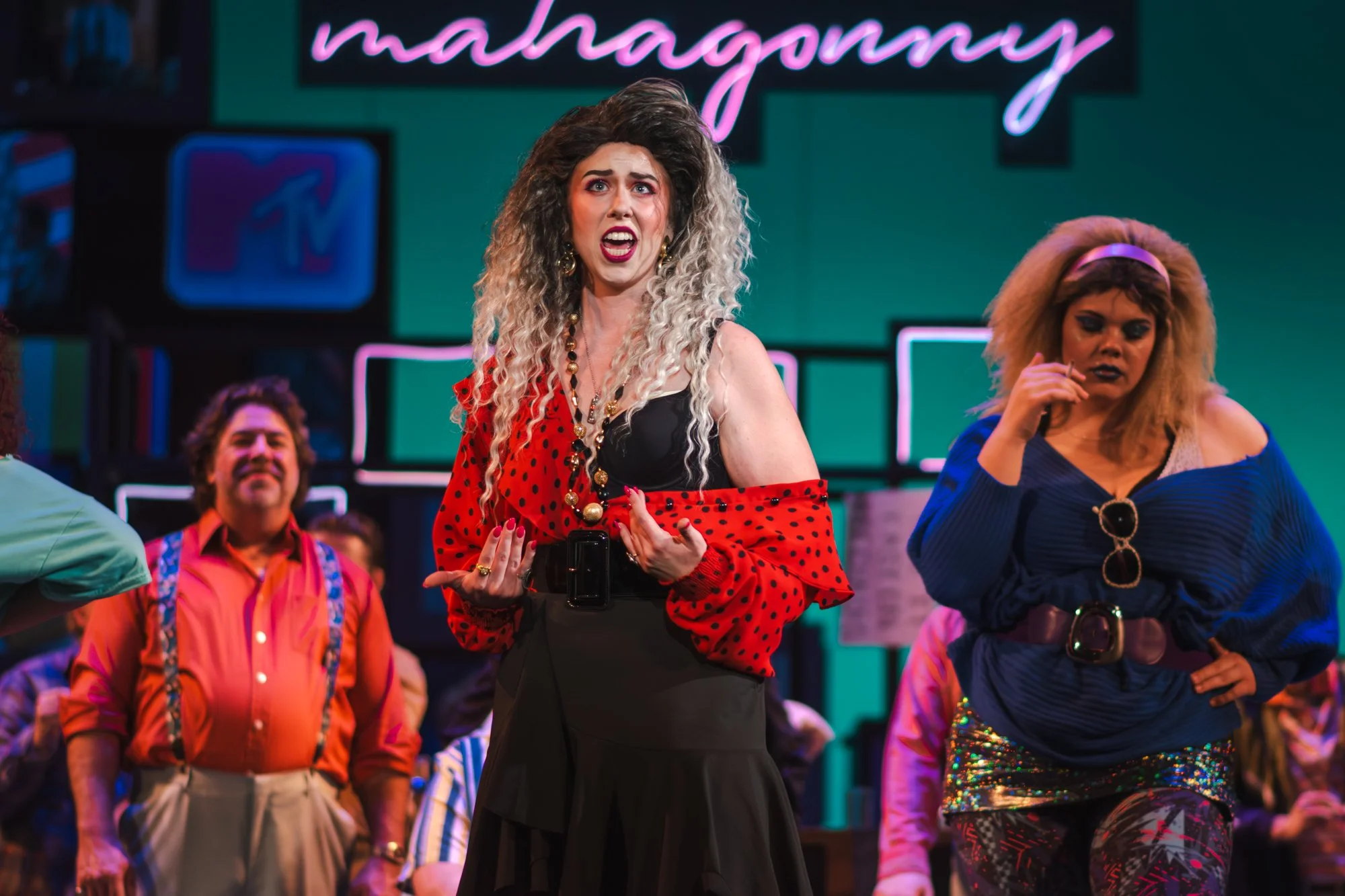 The image shows a theatrical performance with actors on stage. A woman with long curly blonde hair, wearing a red polka dot shirt, black skirt, and bold jewelry, appears to be speaking or singing passionately. Behind her, one actor is smiling, and an