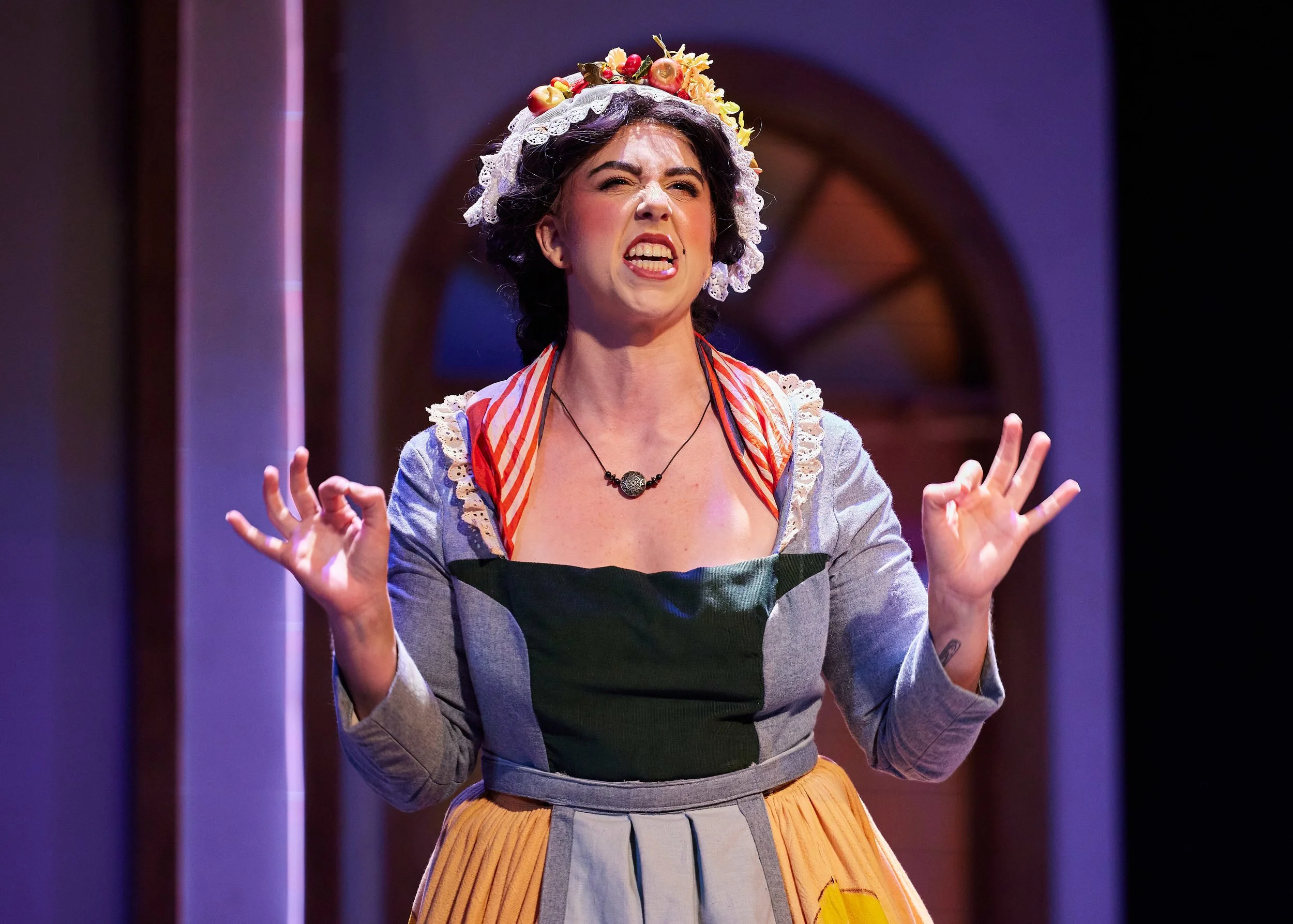 A woman performing on stage, dressed in a period costume with a floral headdress, gesturing with her hands and making an expressive facial expression.